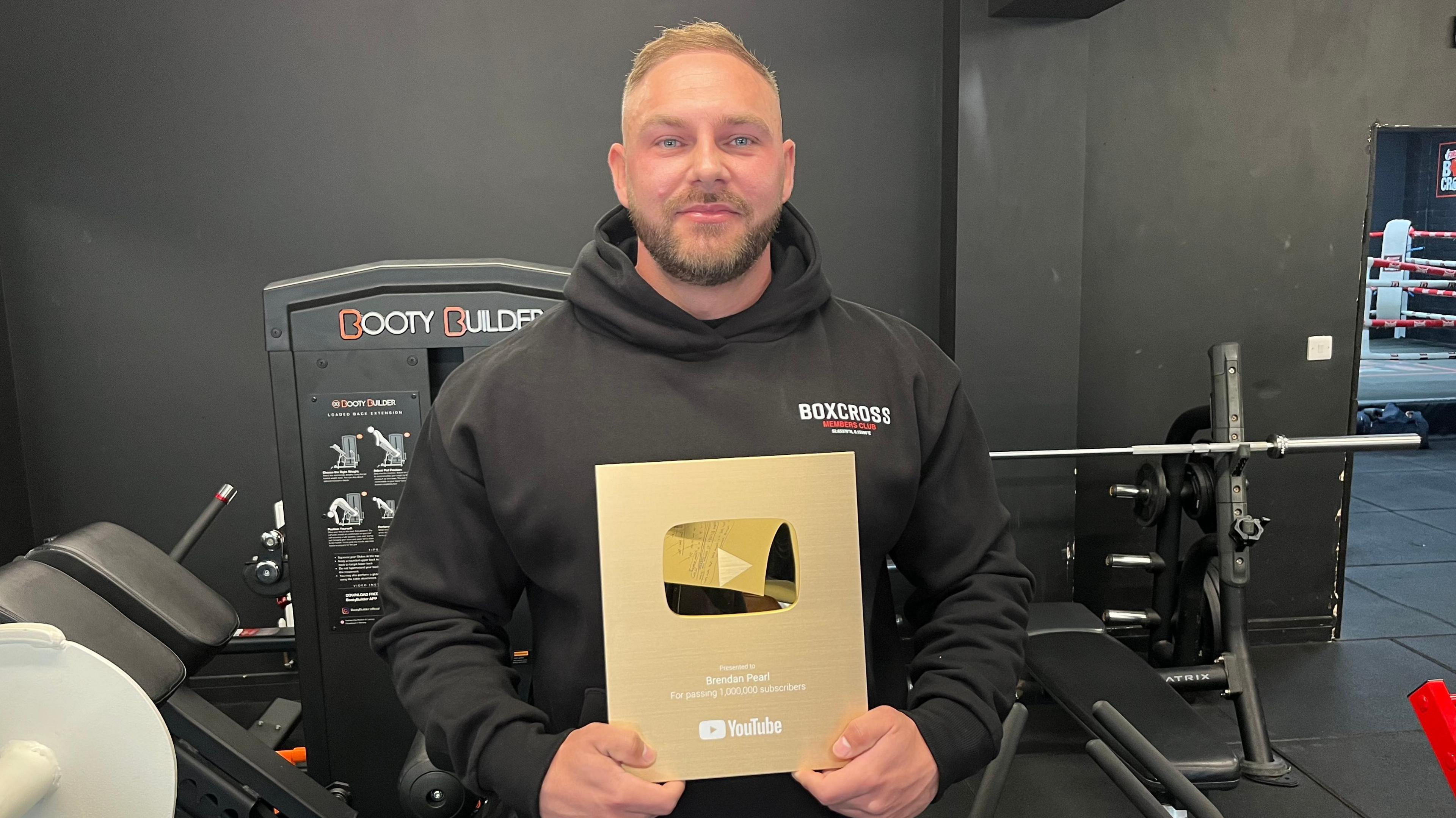 Brendan Pearl with short dark-brown hair with a quiff at the top. He has a matching neatly trimmed beard and a moustache. He is wearing a black tracksuit top with the hood down. Behind is a black wall with various gym equipment visible. He is holding a gold plaque from YouTube, which marks the fact he has one million subscribers.