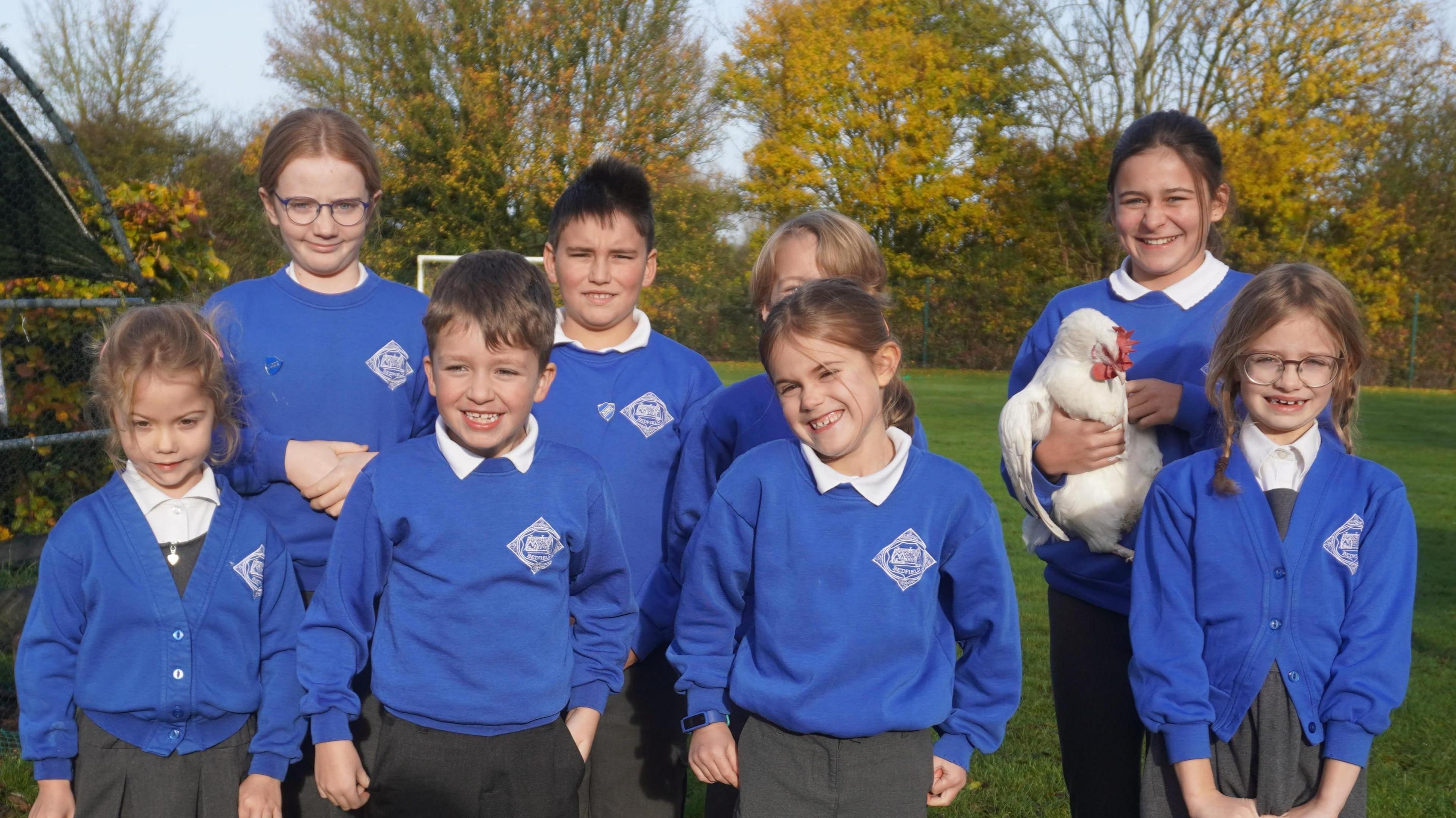 A small group of pupils stand in a group to pose for a photo. Taller and older students stand in a line behind another line of smaller younger students. One of the older students is holding a white hen. They are all wearing blue school jumpers with white shirts underneath.