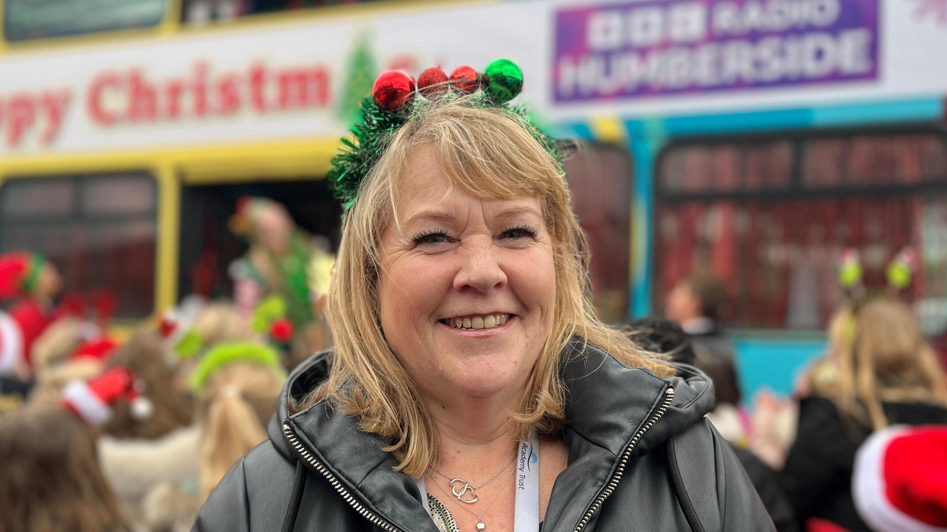 Joanne Mulligan. She has light brown hair and is wearing a black jacket. She has green tinsel on her head with four baubles - three of them red and one of them green. The BBC Radio Humberside Santa Bus is in the background with a crowd of children facing it. 