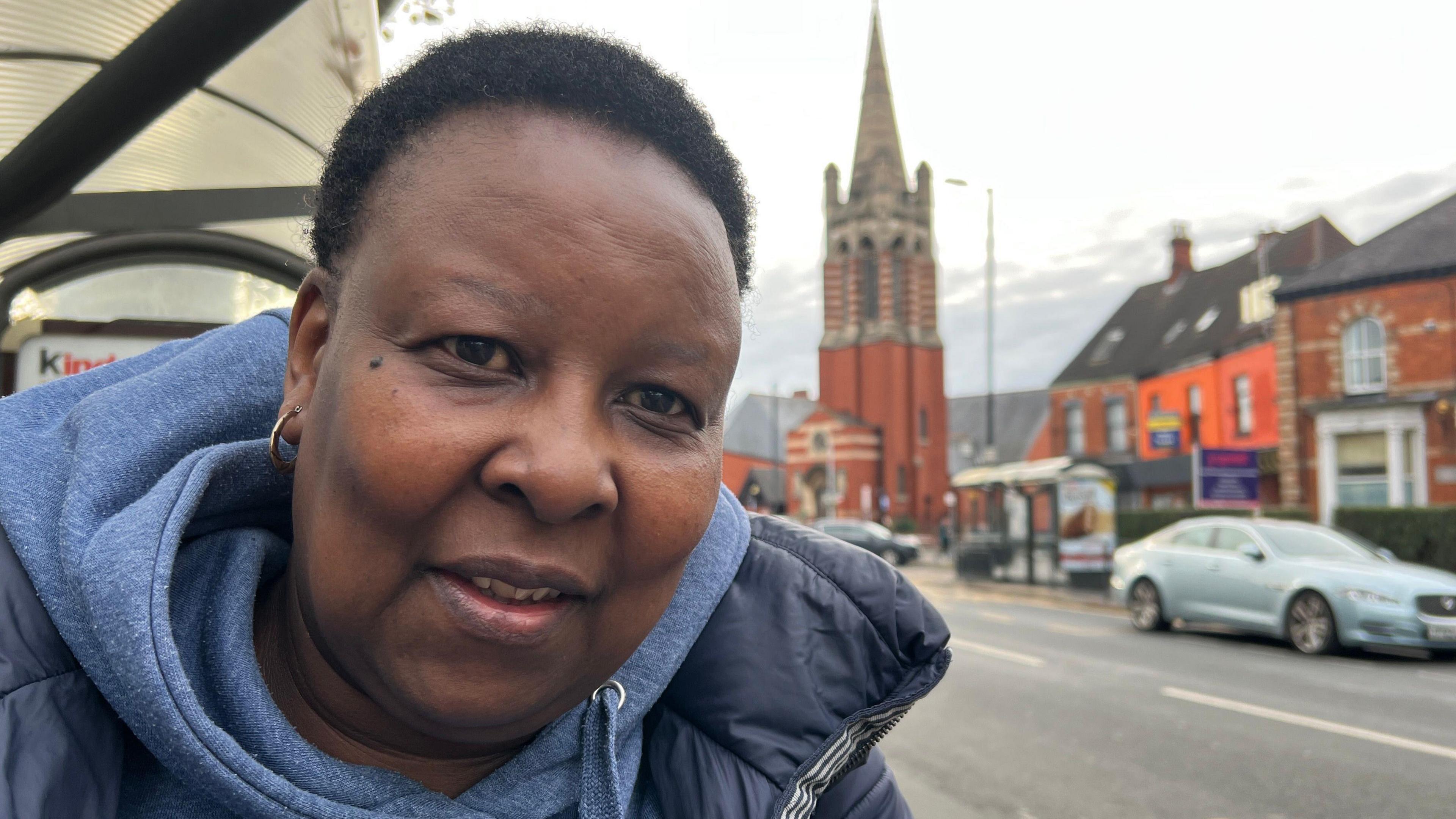 A woman wearing a blue winter coat and hoodie stands at a bus stop. She has short brown hair. A red-brick church and steeple can be seen in the background.