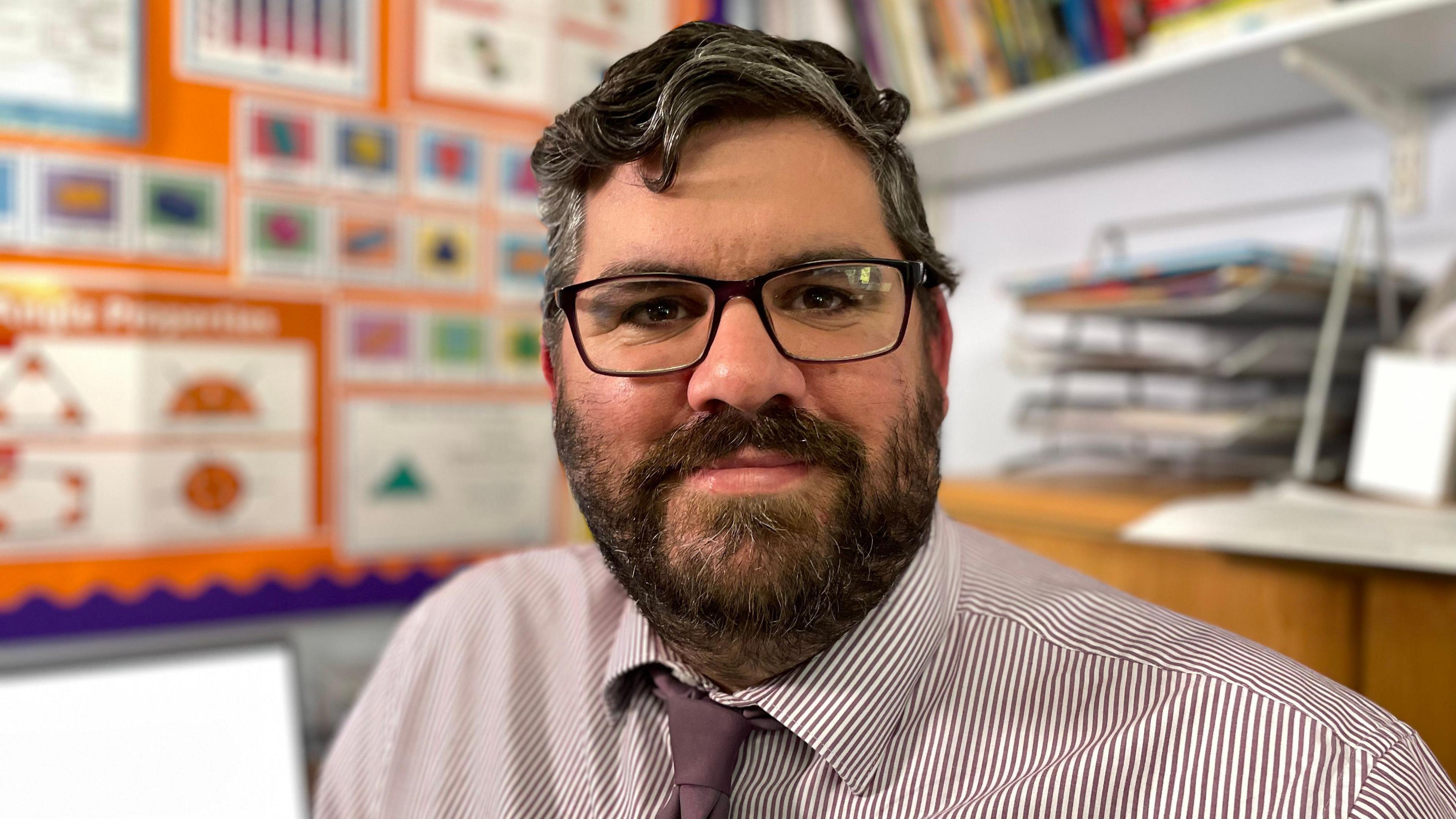 Head teacher in a classroom wearing glasses and a striped shirt. The background display features mathematics.