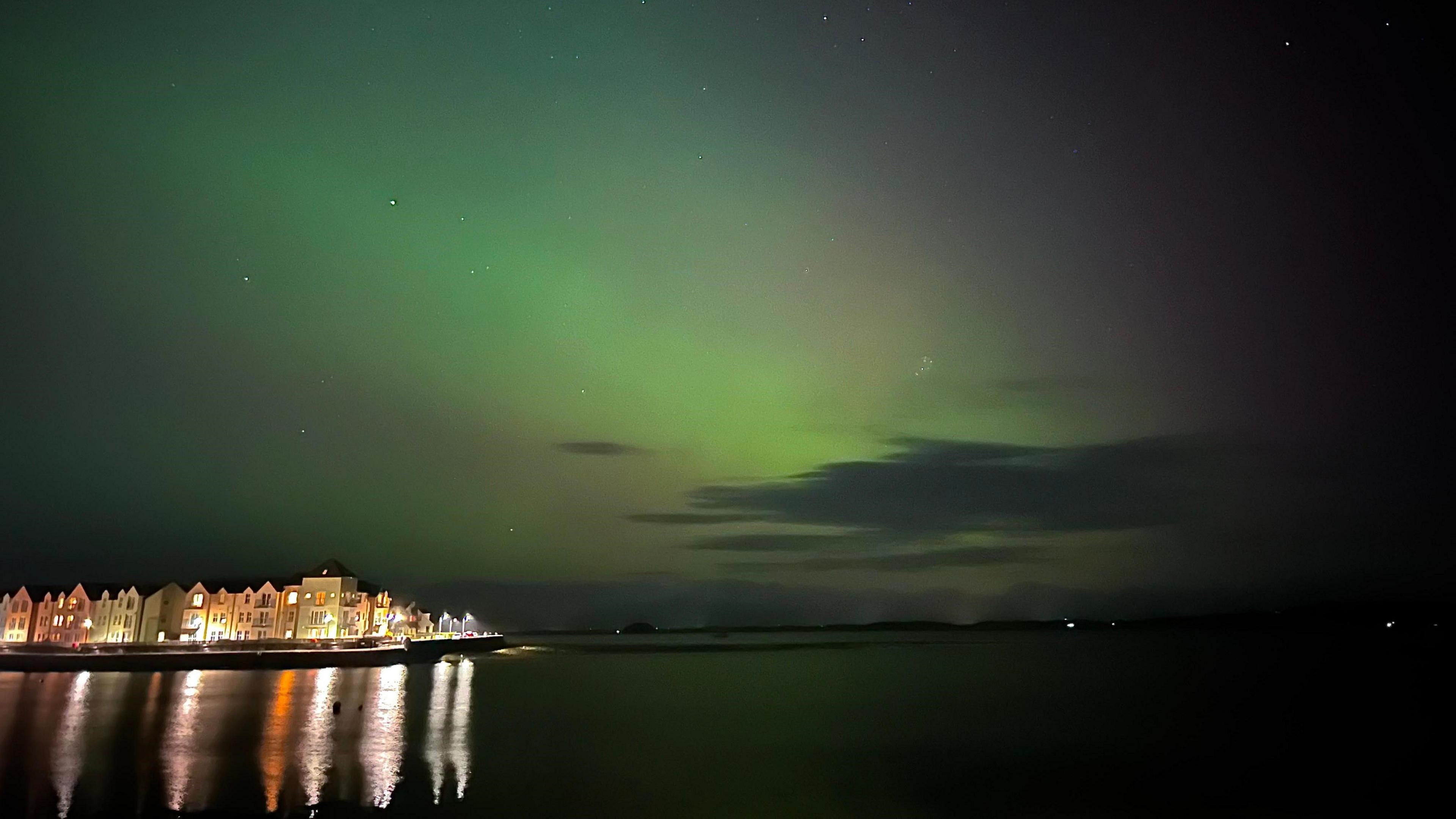 The sky lit up green with Northern Lights. There is a body of water in the bottom half of the image with a row of homes lit up to the left of the water. The reflection of the lights from the homes is visible in the water. 