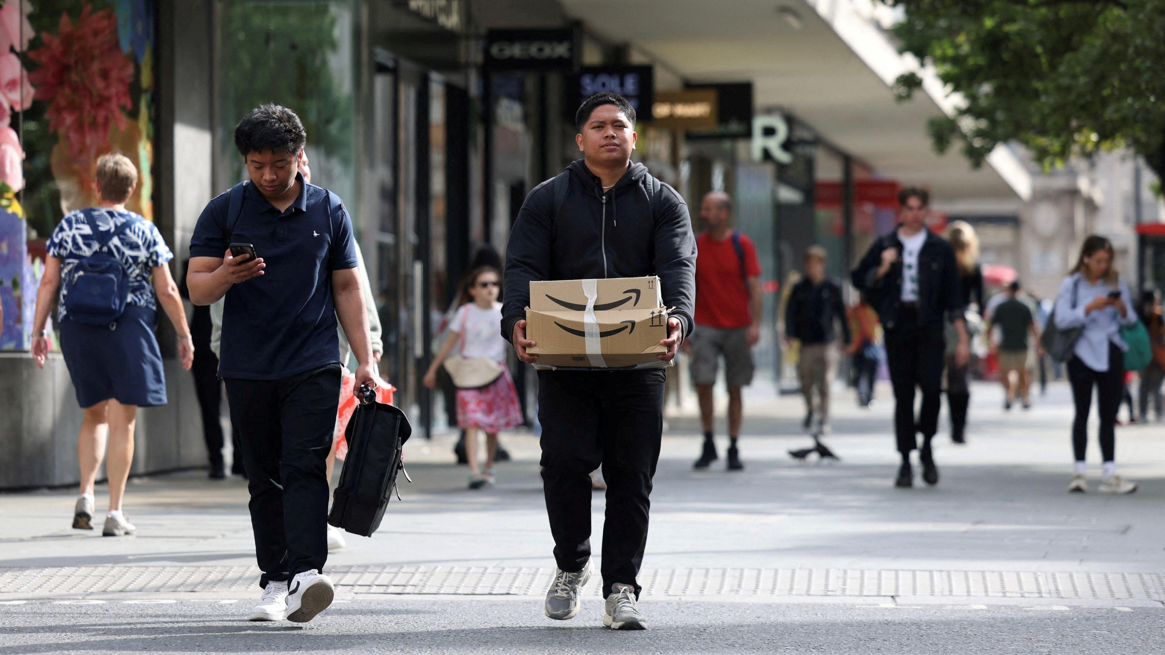 A man walks past shops on Oxford Street carrying two brown boxes with Amazon logos on them. Crowds of people pass in the distance