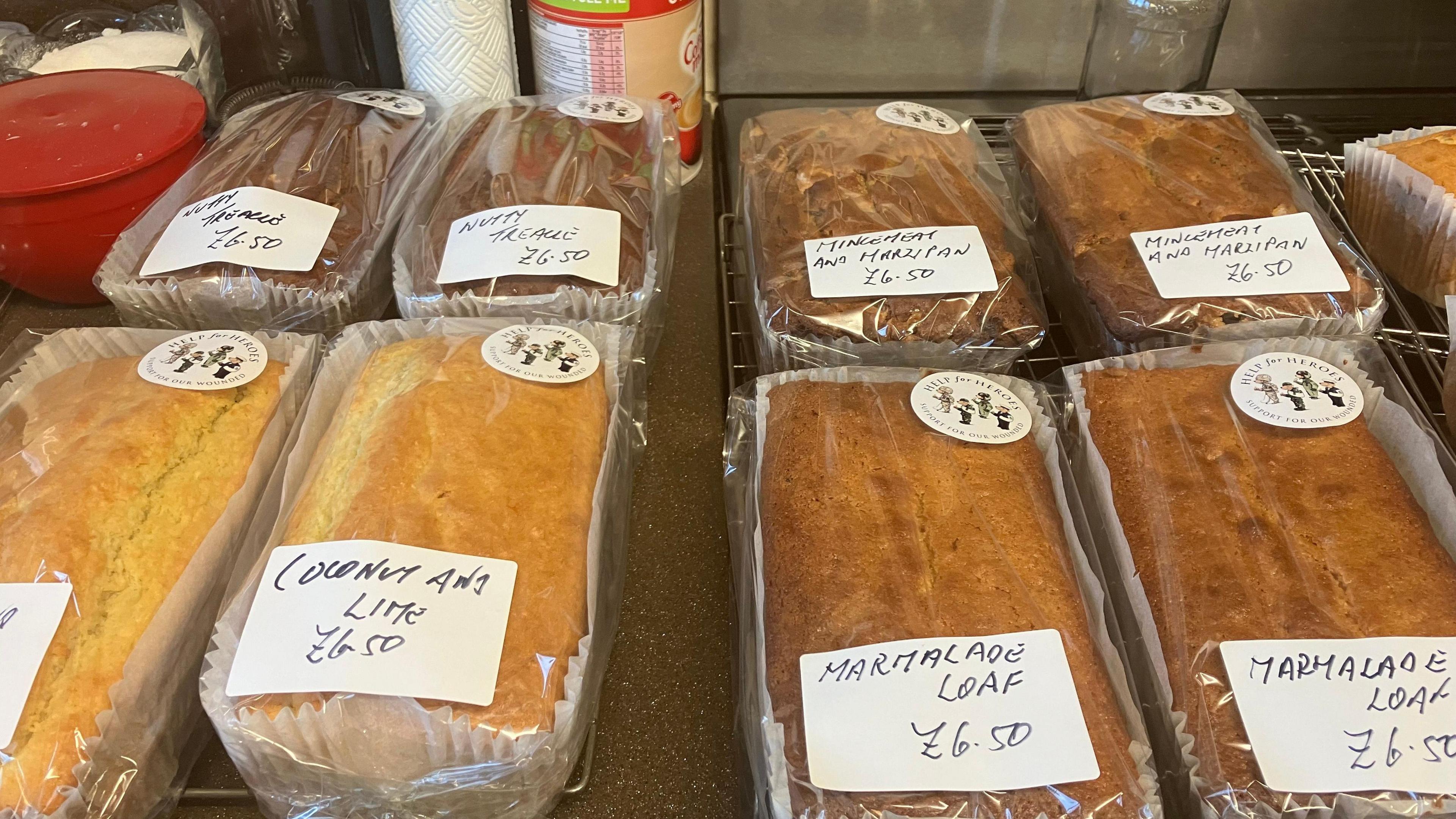 A selection of cakes covered in clear plastic wrapping are on a table with stickers on them reading coconut and lime, nutty treacle and marmalade loaf     