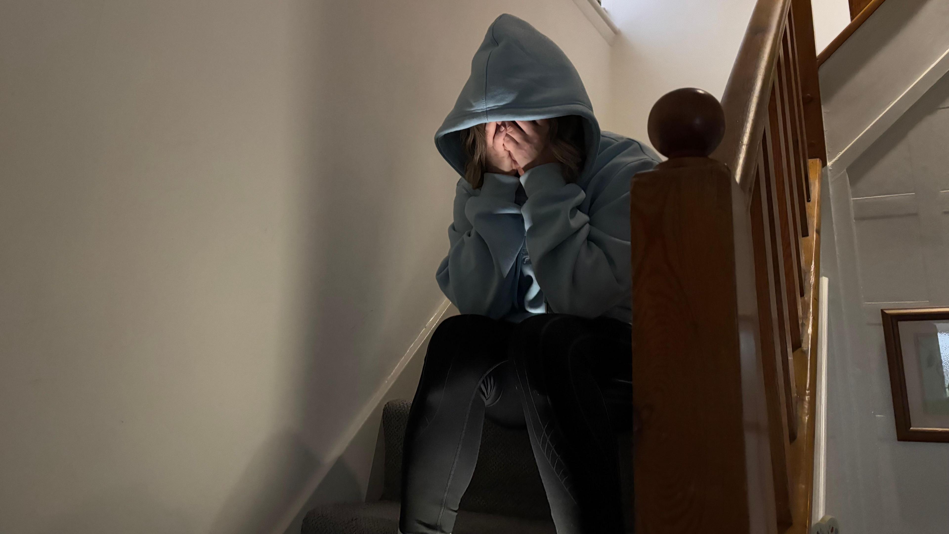 An unidentified woman sitting on a staircase in a house. She is wearing a blue hoodie and black jeans and her face is obscured as she has her head in her hands.