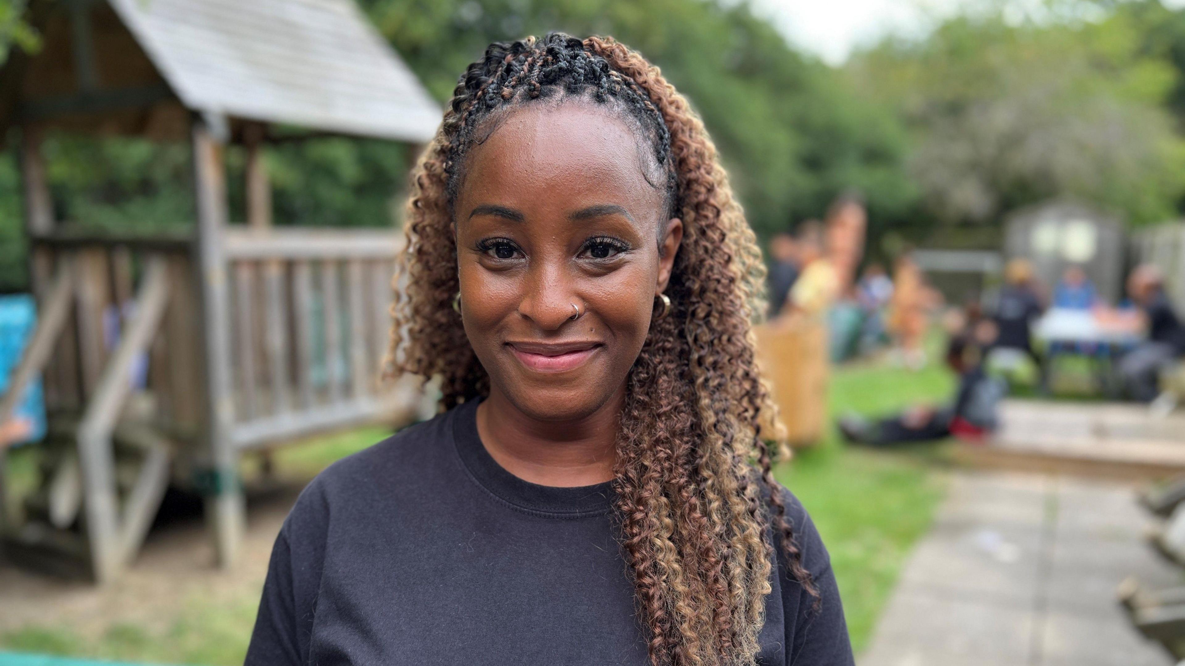 Precious Graham smiling to the camera. She is wearing a black t-shit and her plaited black hair, with blonde tips, is pulled pack in a long pony tail. she is wearing a nose ring on her left side and half hoop, medium chunky earrings. 