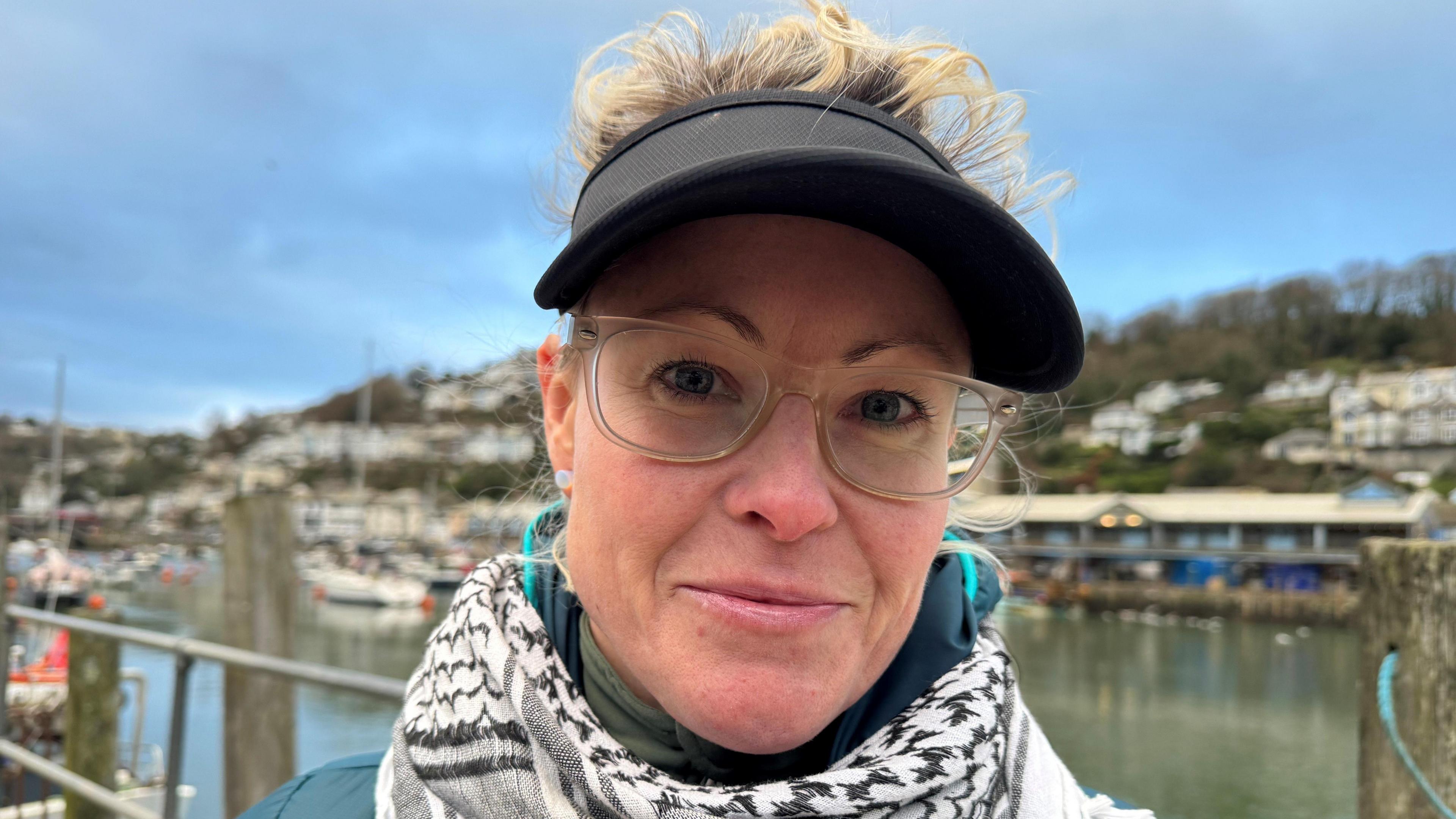 Woman wearing glasses and a visor smiling at the camera with Looe harbour in the background. 