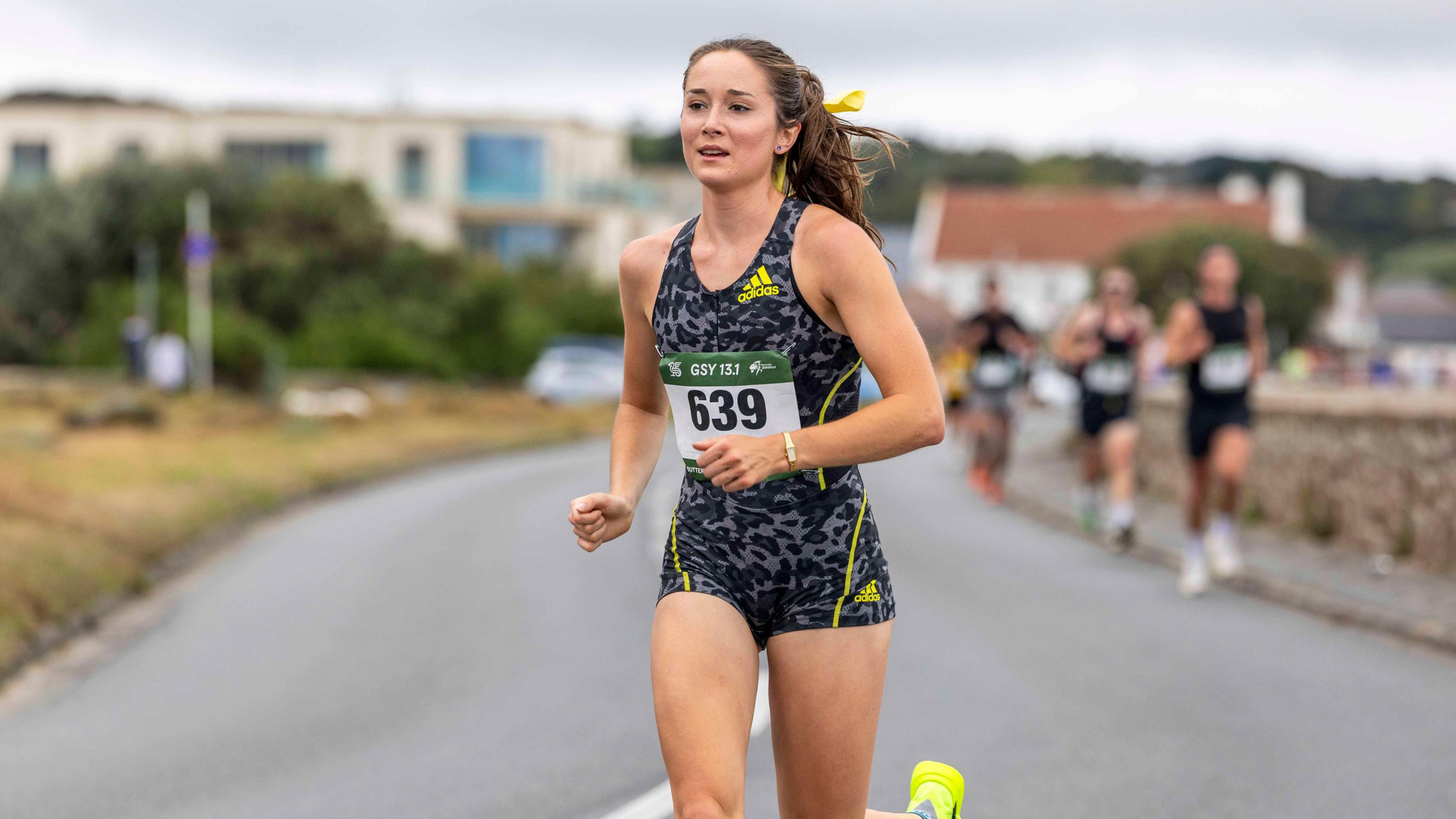 A brunette woman in a grey running outfit and luminous yellow trainers runs along a costal road, with several runners behind her in the distance.