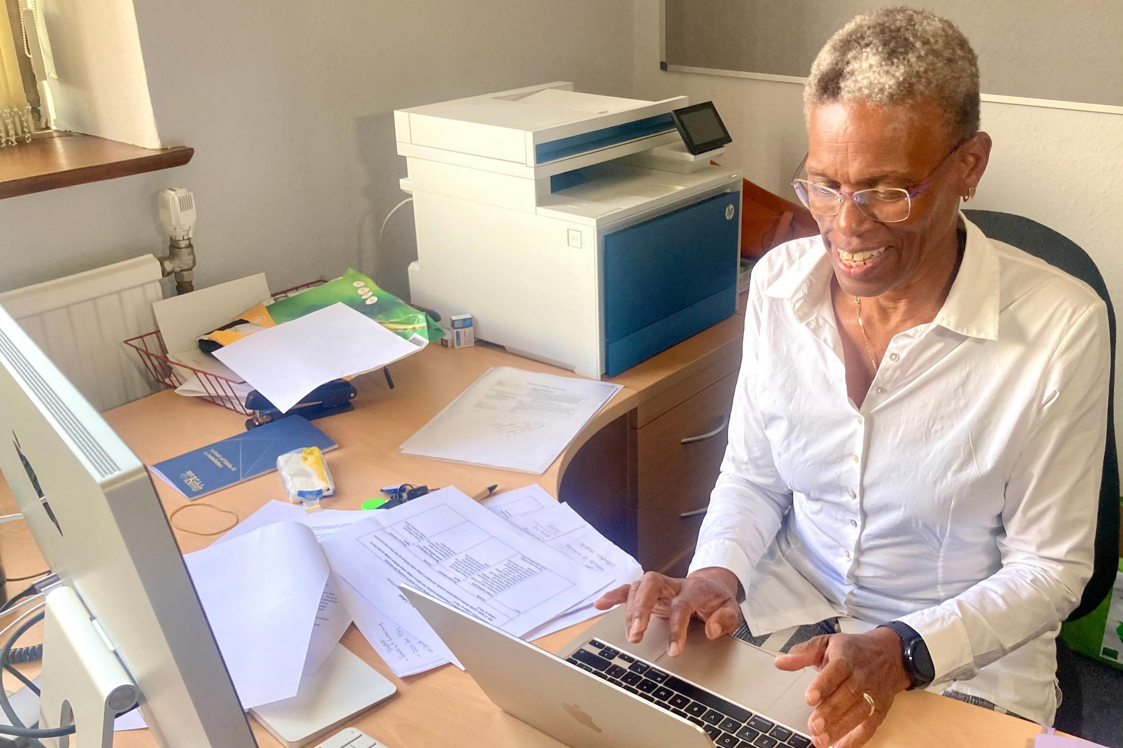 Ijeoma Uchegbu wearing a white shirt, leaning over a desk and a laptop. She is wearing glasses, has short hair and is smiling.