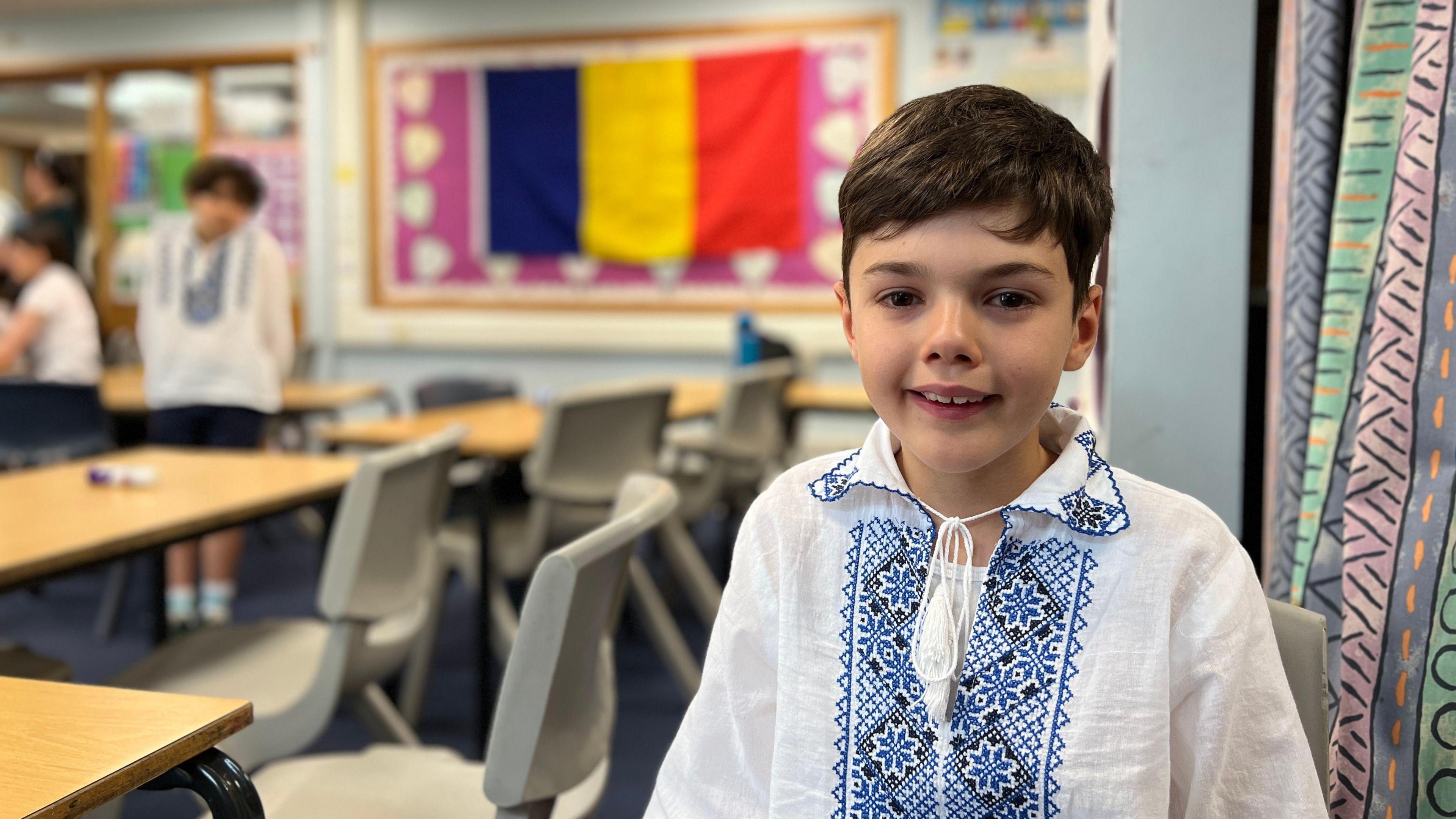 Classroom with students wearing traditional embroidered shirts. Stefan is in the foreground. In the background, other students are seated and a large flag with vertical blue, yellow, and red stripes hangs on the wall. Desks and chairs are arranged in rows