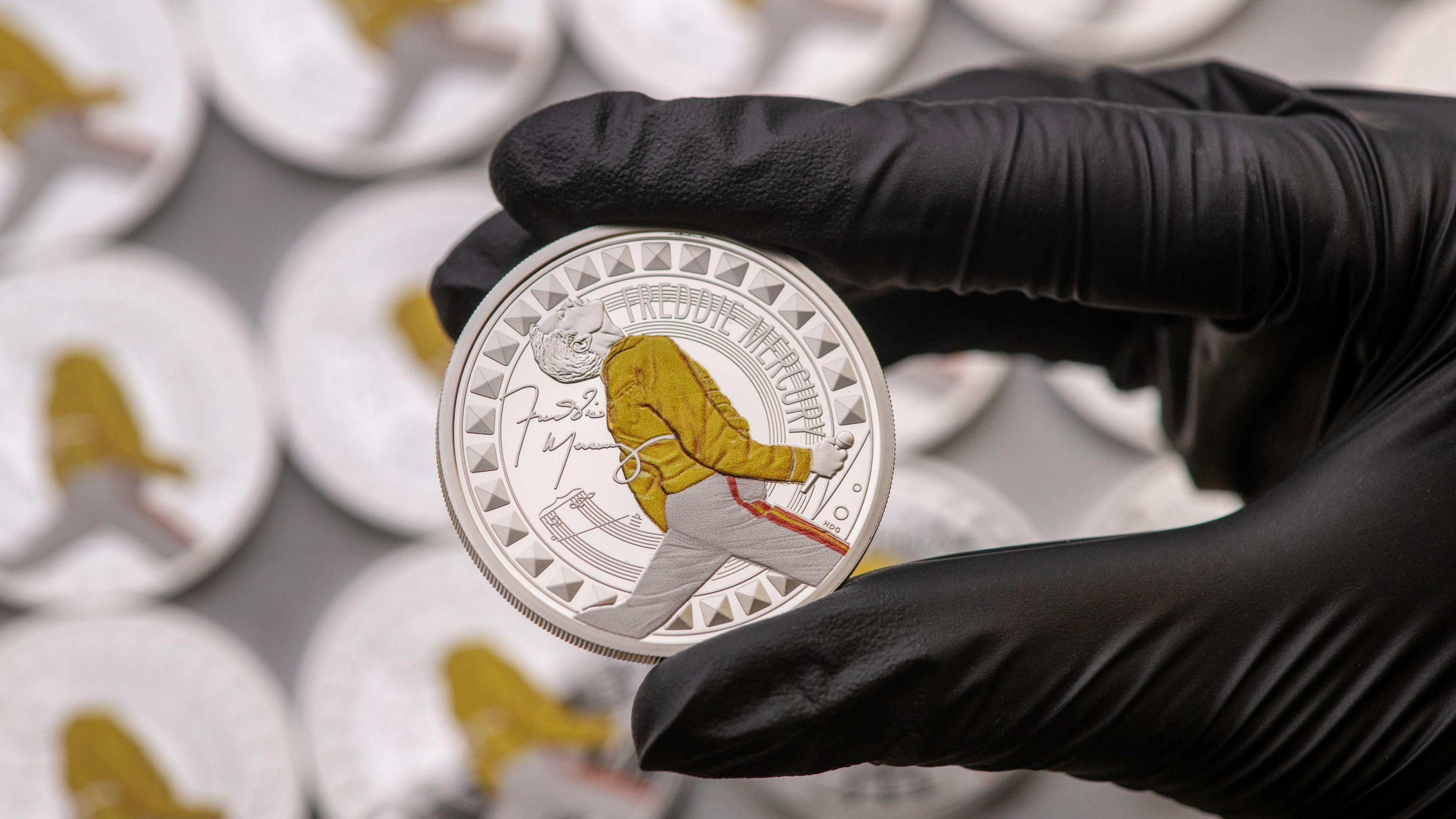 A black gloved hand holds the Freddie Mercury coin. The coin is silver and has an image of Freddie Mercury on the front, mid performance, with his head titled back. The only colour on the coin is his yellow jacket and white trousers. The coin also has Freddie Mercury's signature on the left hand side. 