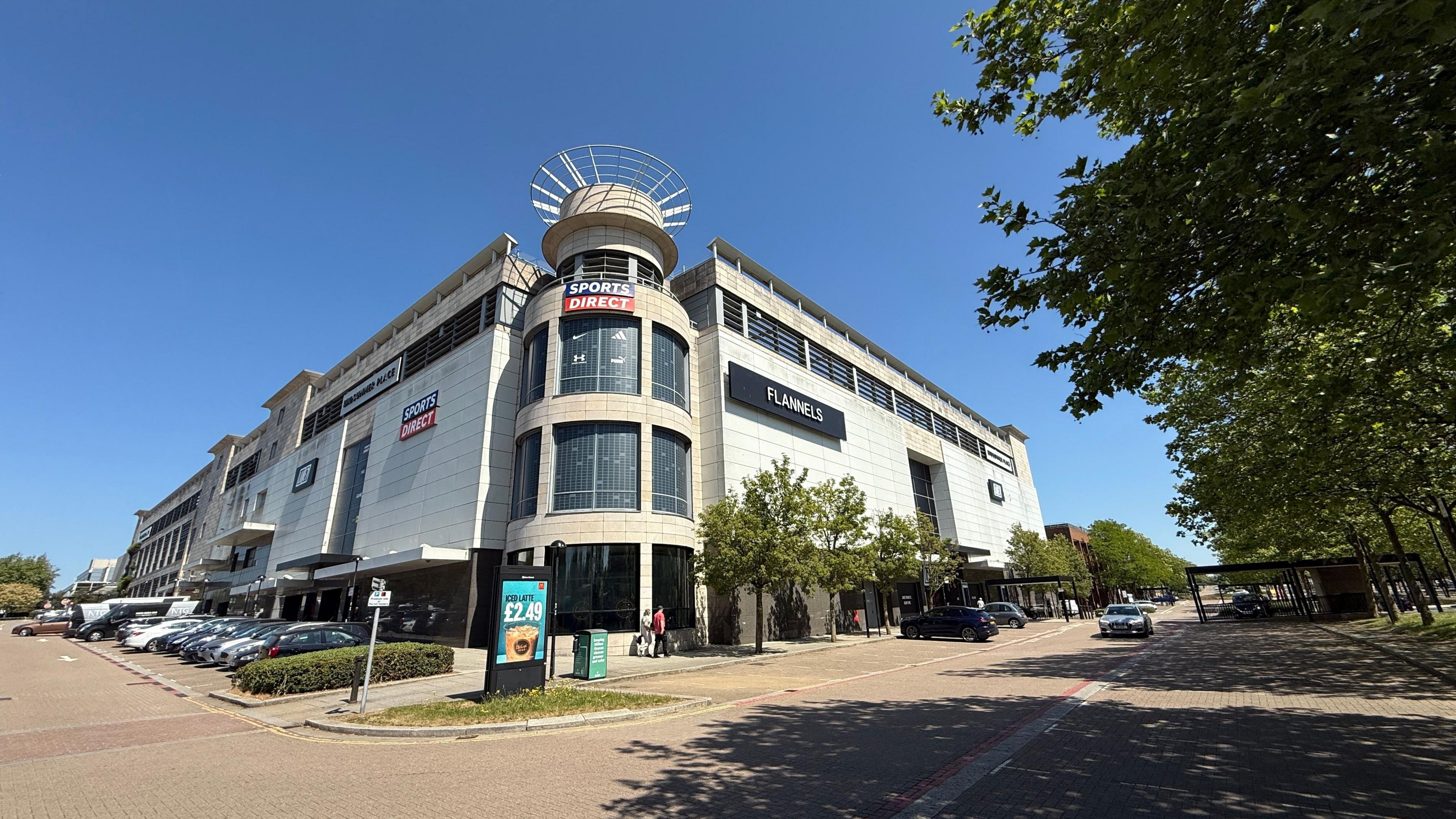 The exterior of the former Debenhams in Milton Keynes, with a large turret-shaped building in the middle surrounded by two large rectangular buildings. The sides of the buildings say Sports Direct and Flannels. In the foreground is a car park.