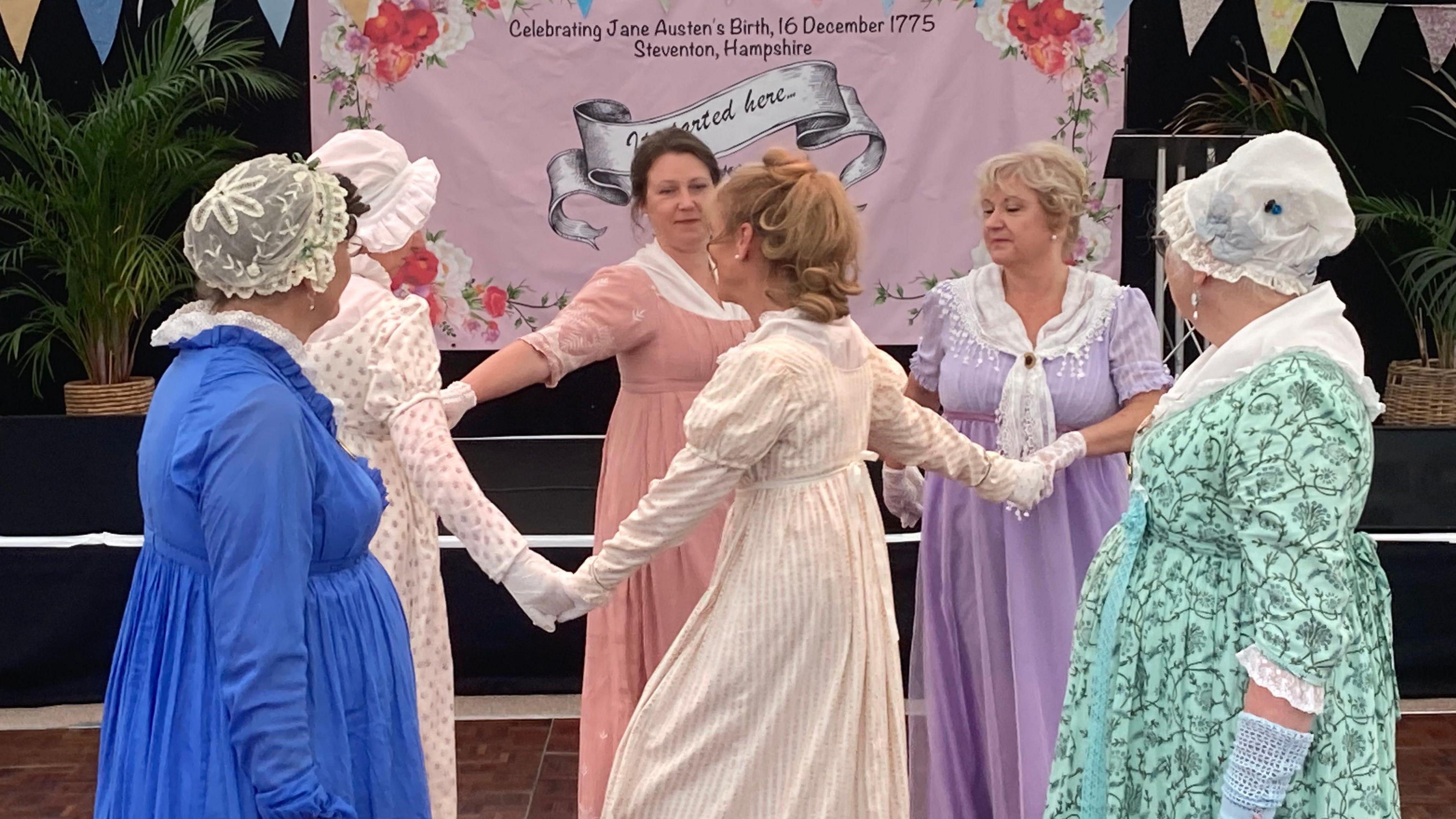 A group of women holding hands and dancing in a circle dressed in Regency regalia.