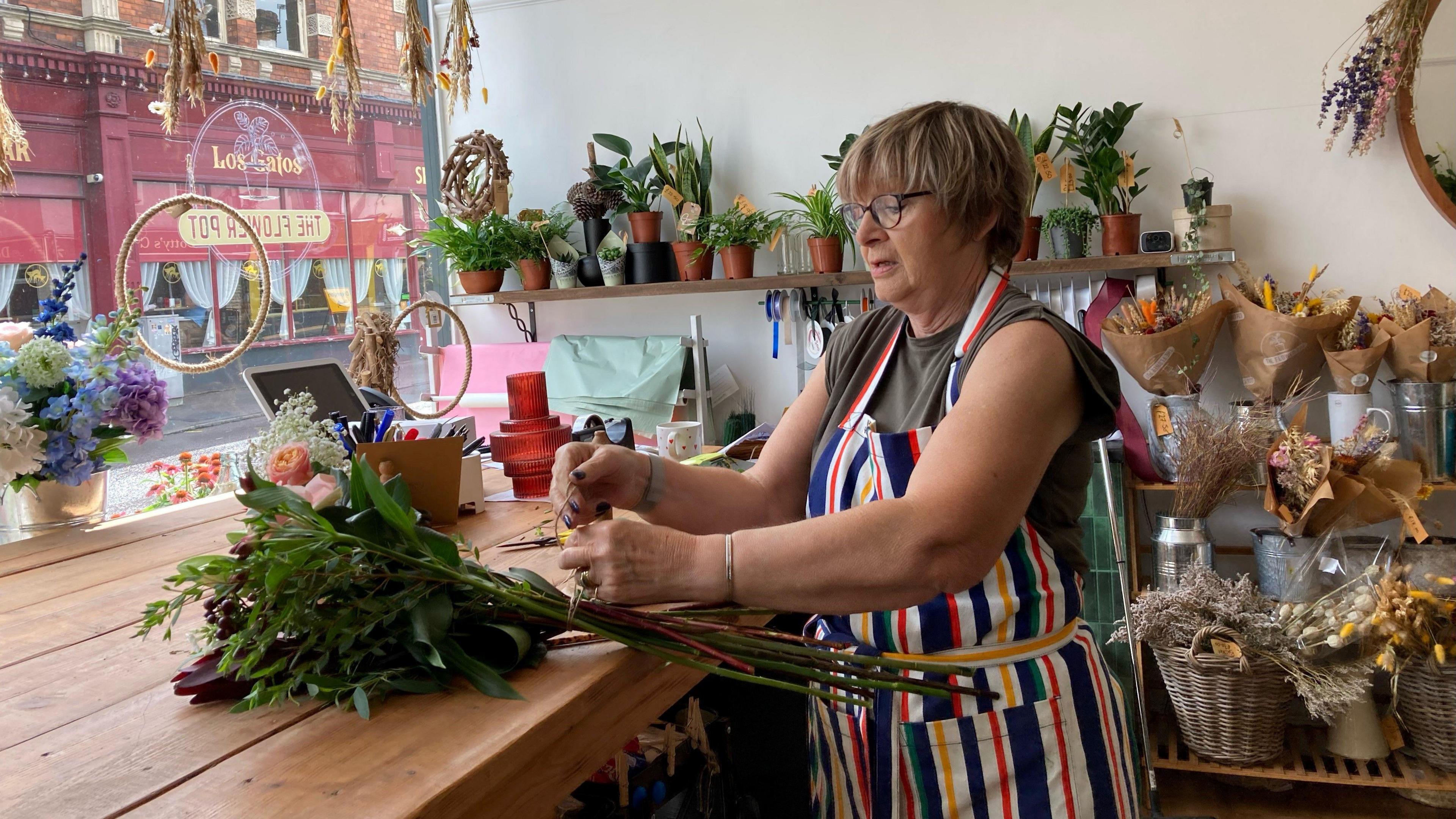 Flower Pot owner preparing bouquet in her shop. She is wearing a stripy apron and there are plant pots on shelves in the background.
