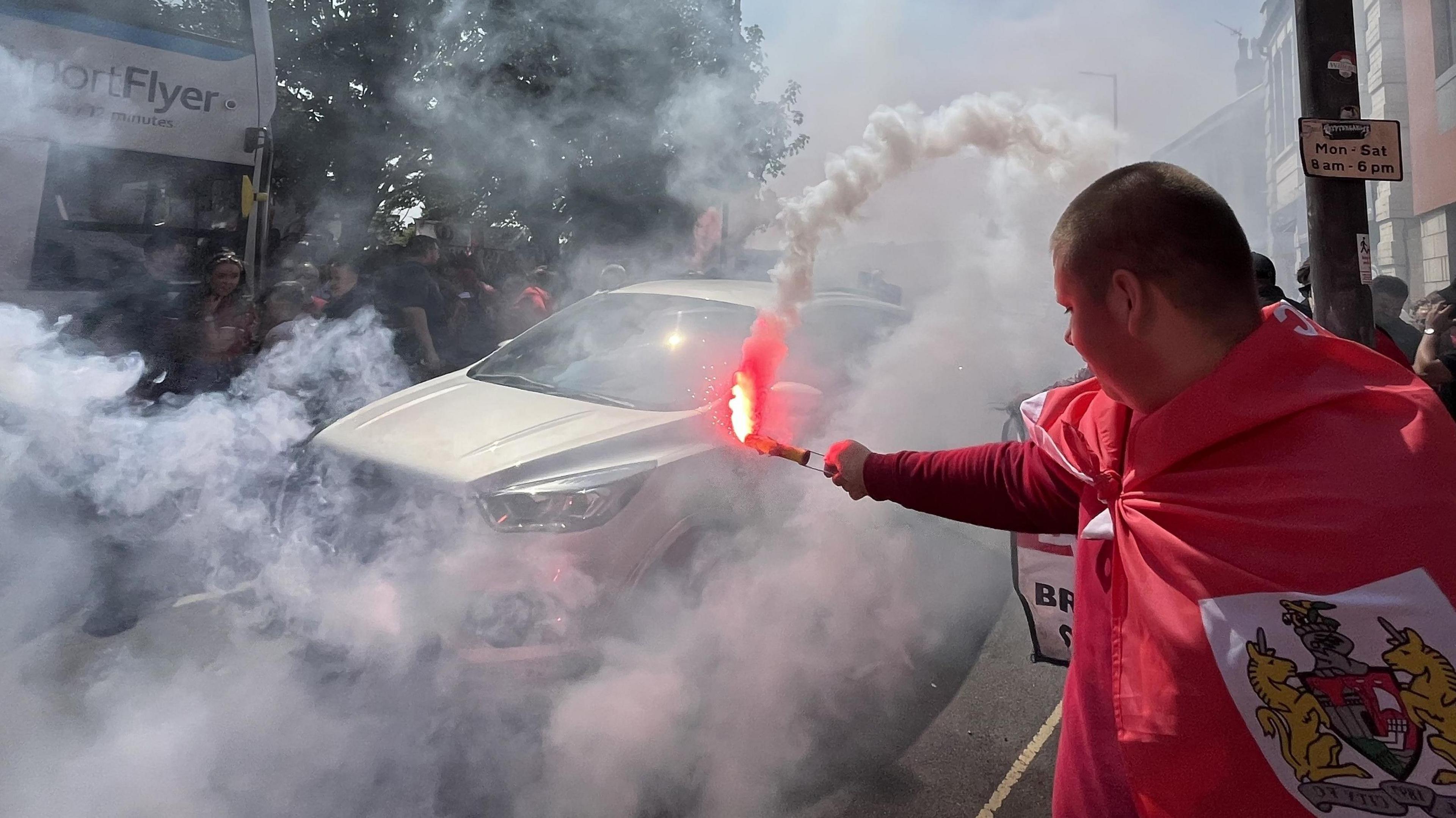 A man with short-cropped hair, draped in a red and white Bristol City flag, holds a smoke flare out as he stands on the pavement as part of the annual Flag Day event. A car can be seen negotiating its way through the smoke, as can an A1 Airport Flyer Bus in the opposite carriageway