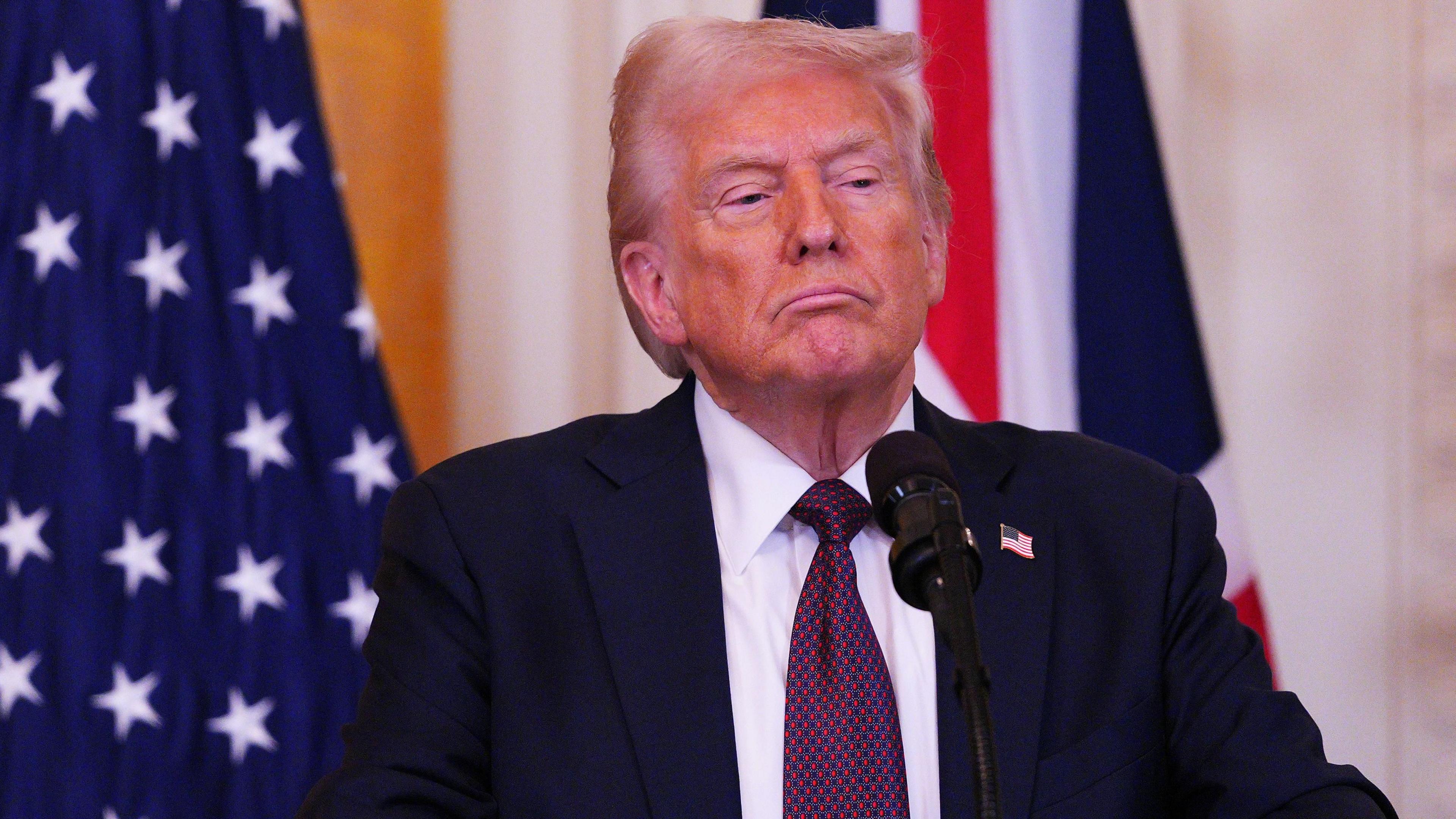 Donald Trump in front of US and UK flags, wearing a dark suit, white shirt and red-and-blue tie