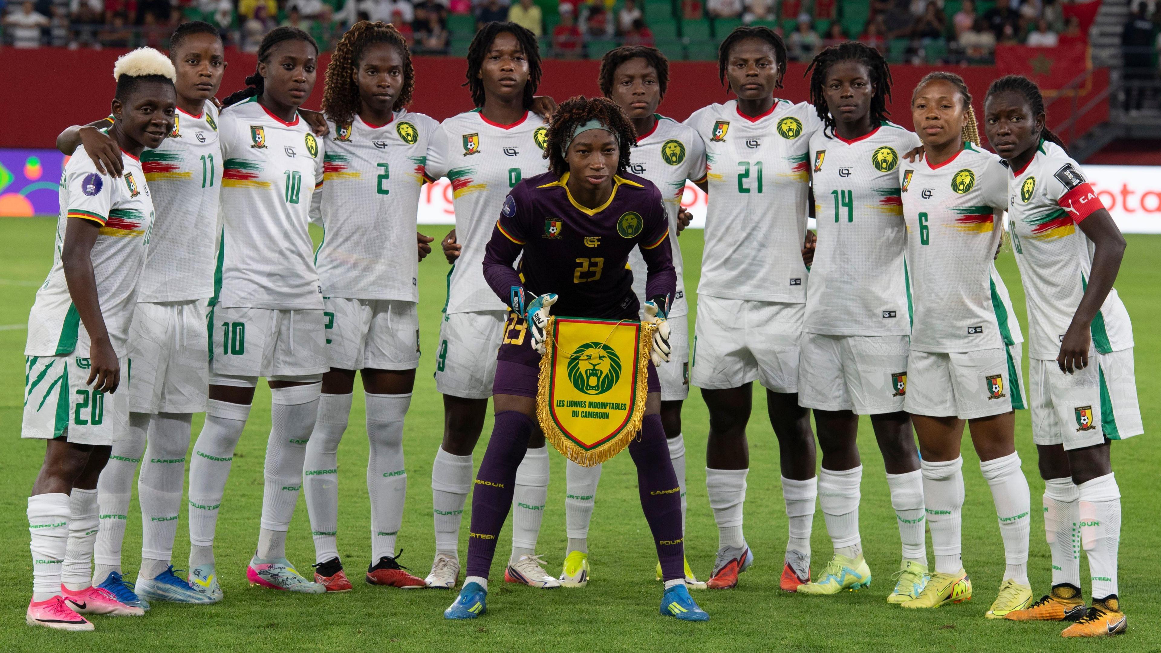 Cameroon's players line up on the pitch for a team photo ahead of a Women's Africa Cup of Nations game. Ten outfield players wear an all-white kit and form a semi-circle around goalkeeper Michaely Bihina, who wears an all-purple kit and holds a yellow Cameroon pennant in front of her