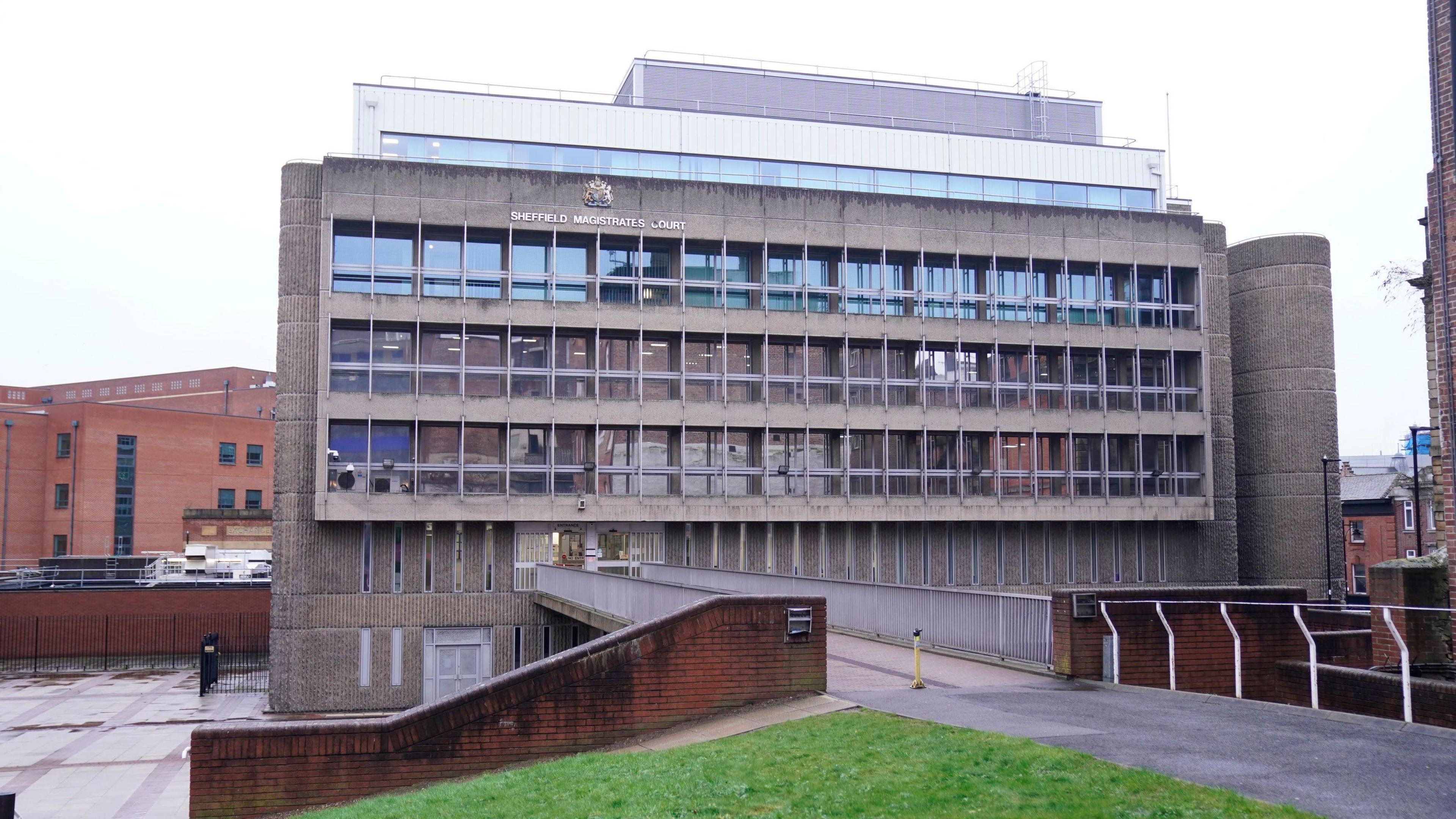 A grey brutalist building, pictured from the other side of a footbridge which leads to it. The building has lots of windows. It's a cloudy day.