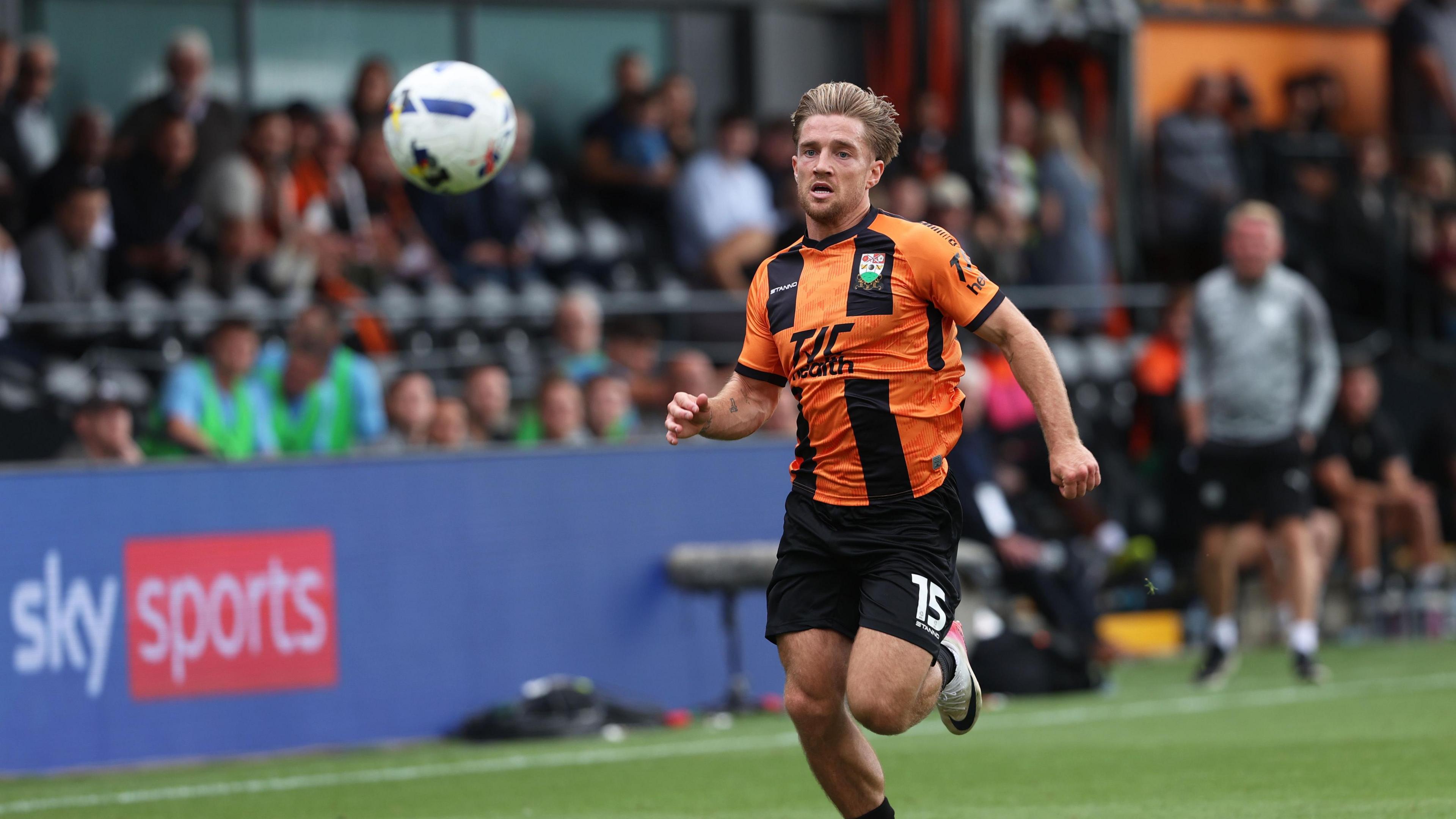 A Barnet player chases after the ball during a League Two fixture. He is wearing an orange and black kit with an advertising hoarding, fans and a coach seen standing in the background.