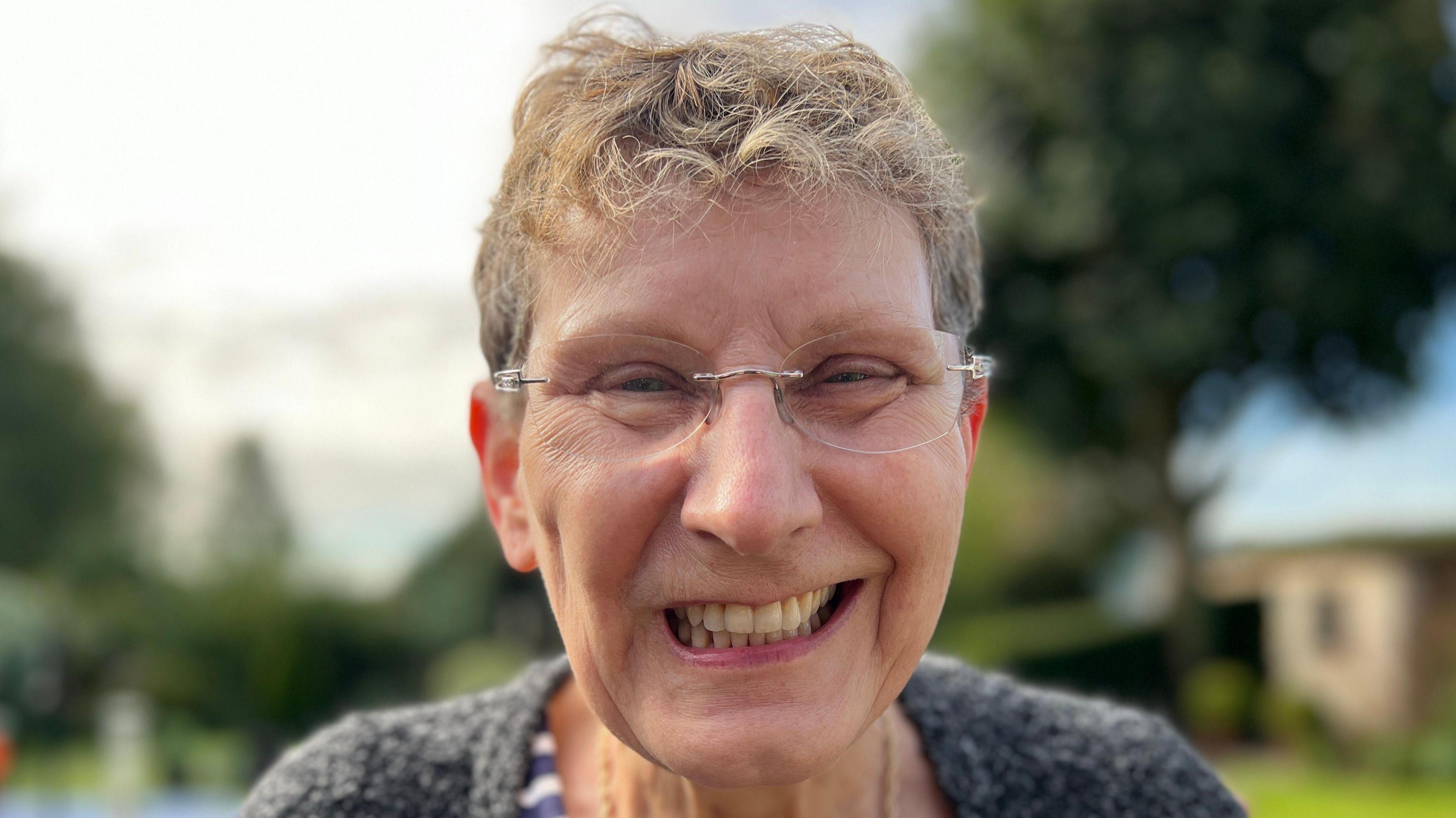 Jane Whittaker has clear glasses which are perched on the apples of her rosy cheeks. Her hair is curly towards the front but short overall with hues of dark brown hidden amongst blonder strands. Jane is standing in front of her garden which has been blurred.