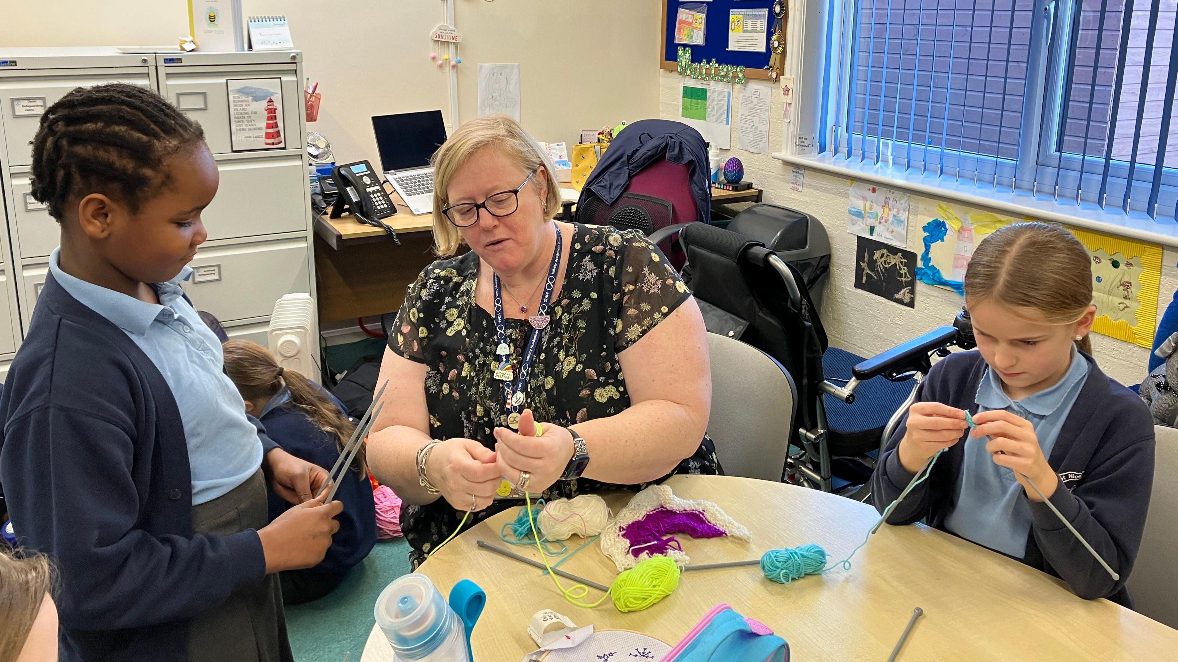 Learning mentor Mrs Curtis is sat at a table in a classroom with two female primary-age pupils. They are all knitting and Mrs Curtis is explaining a stitch to one of the girls. 