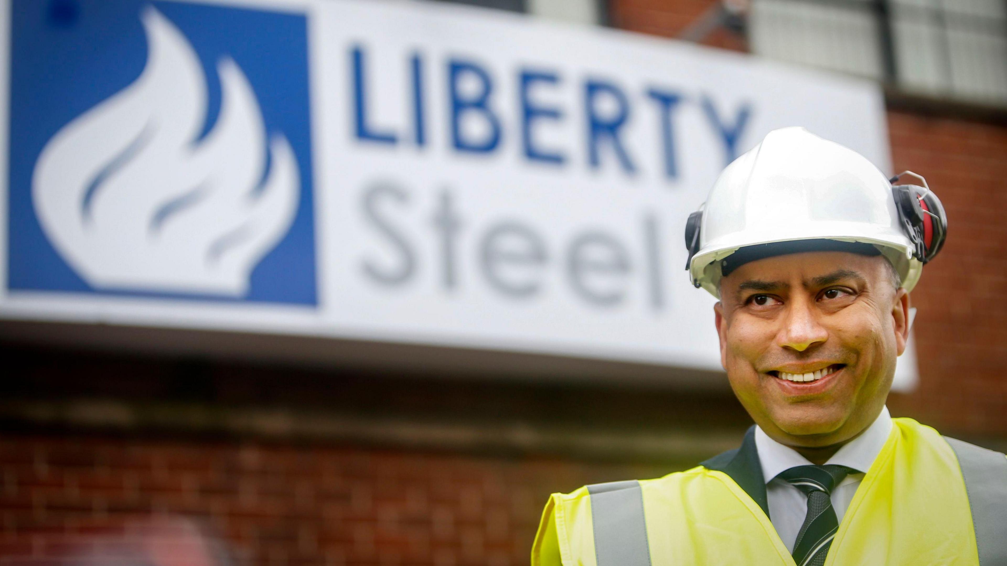 A man in hi-vis and a helmet stands in front of a brick wall with a big sign attached. The blue and white sign reads "Liberty Steel" and features a logo on the left.
