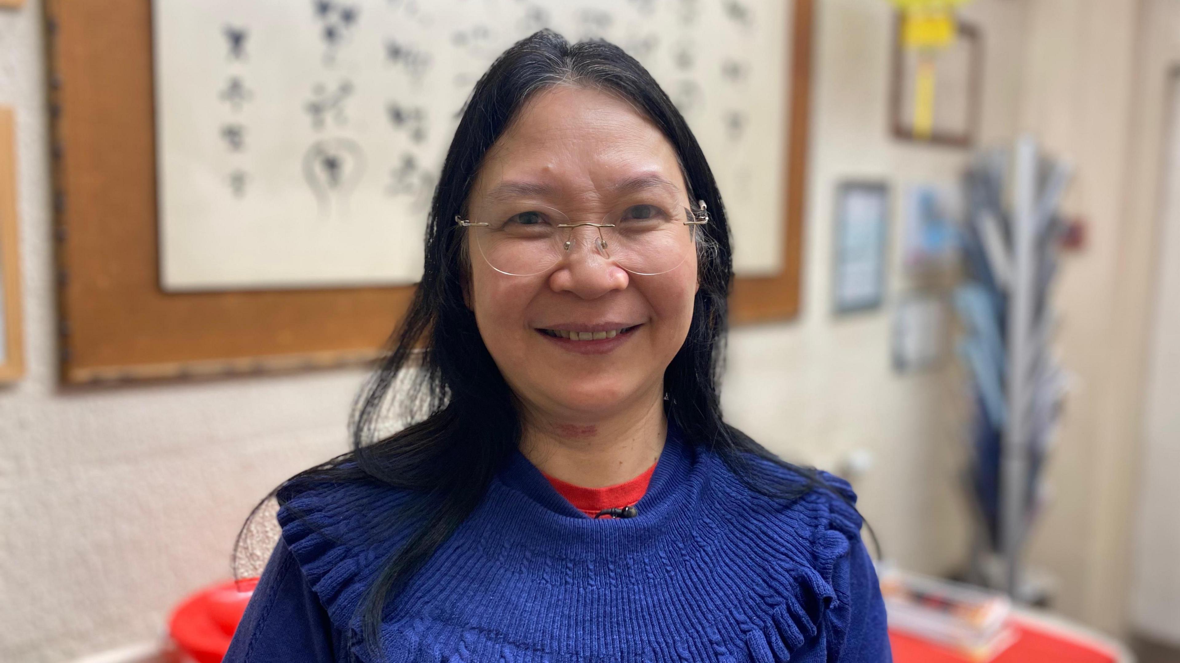 Zi Lan Liao, with dark hair, wears glasses, and a blue jumper and red top, and smiles at the camera 