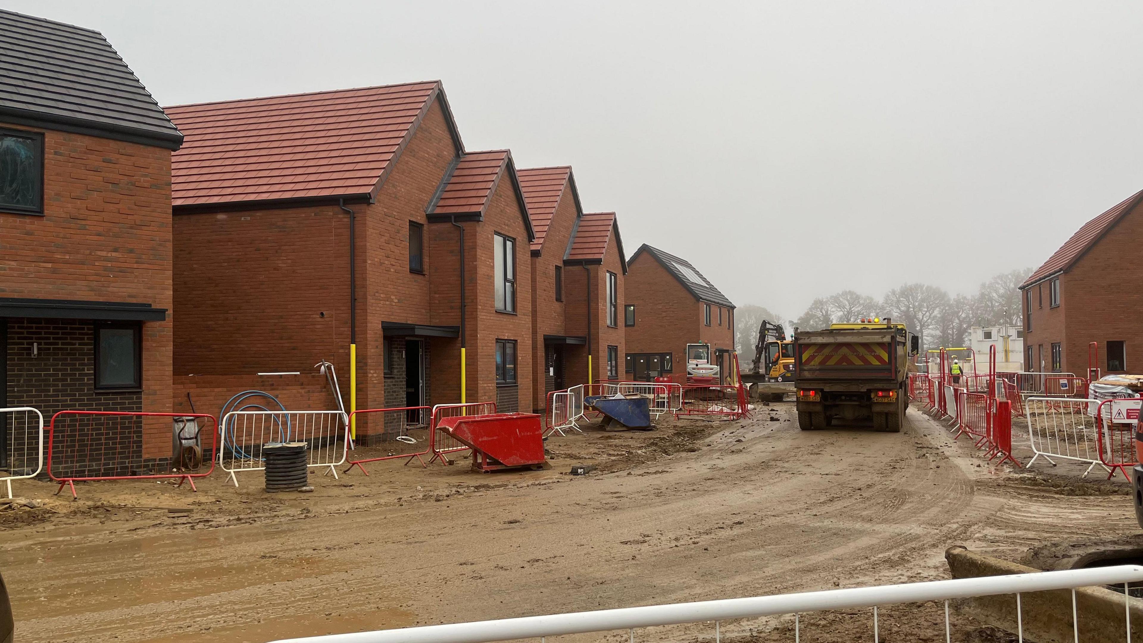 A row of unfinished orange-coloured brick homes on a building site. Machinery and railings can be seen on a dirt-track. 