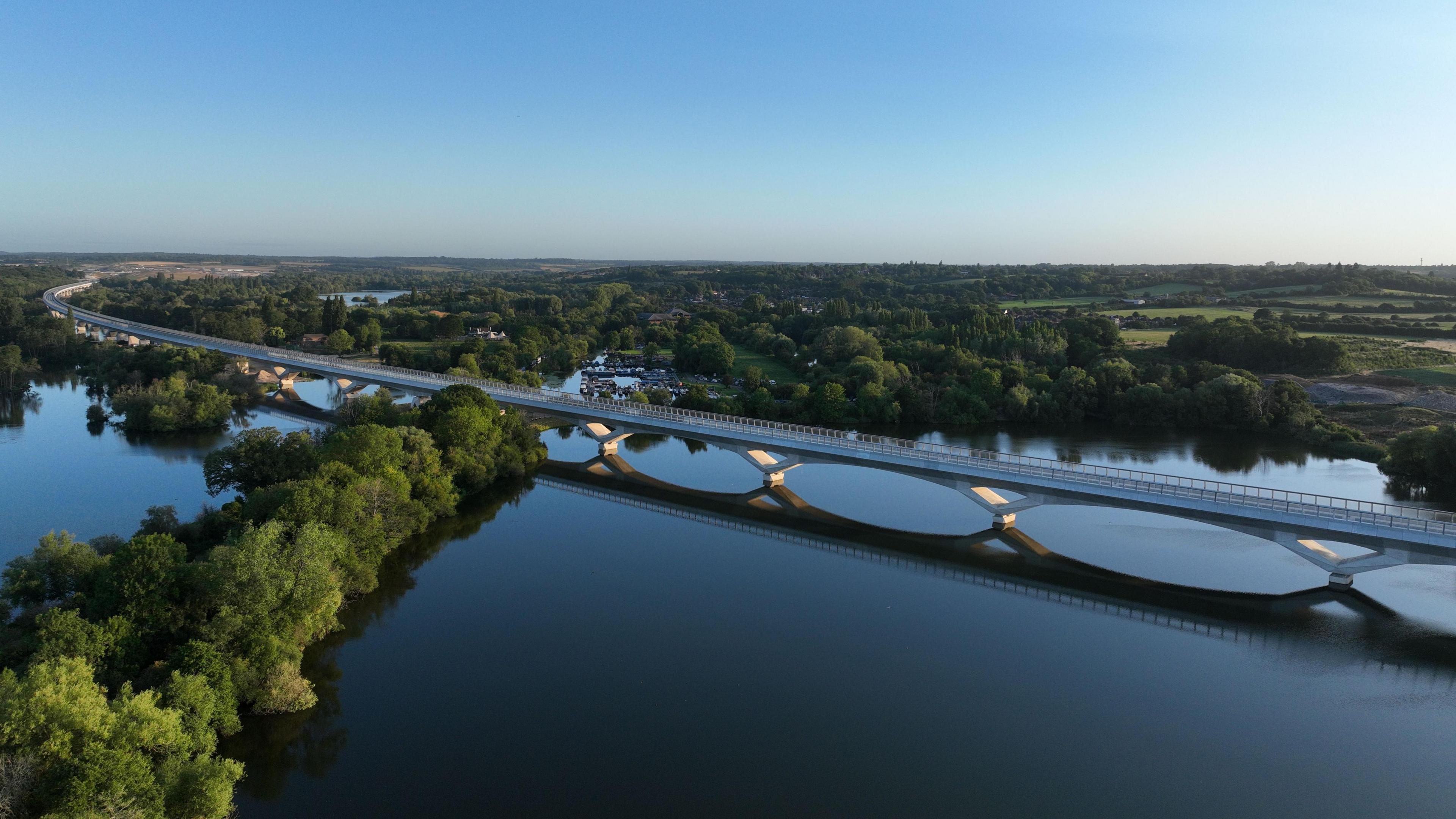A long bridge is going over some water and curves towards the left of the image. There are trees and fields behind the bridge in the background.