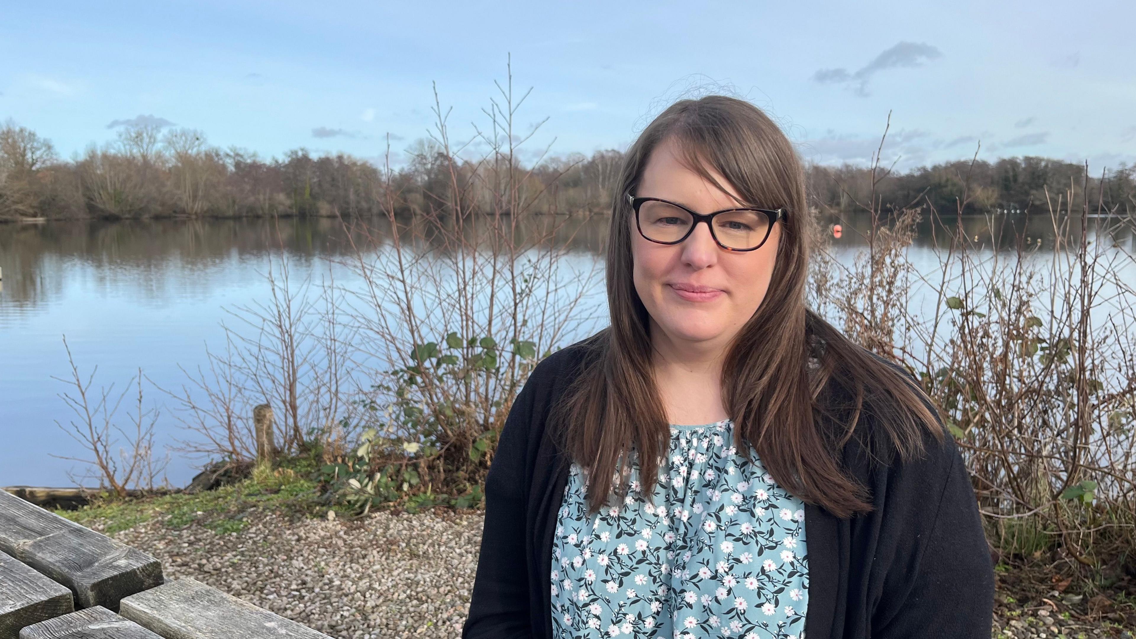 Hayley Jarvis, a 37-year-old woman with long brown hair and wearing a black cardigan, sits on a bench in front of a lake staring directly at the camera