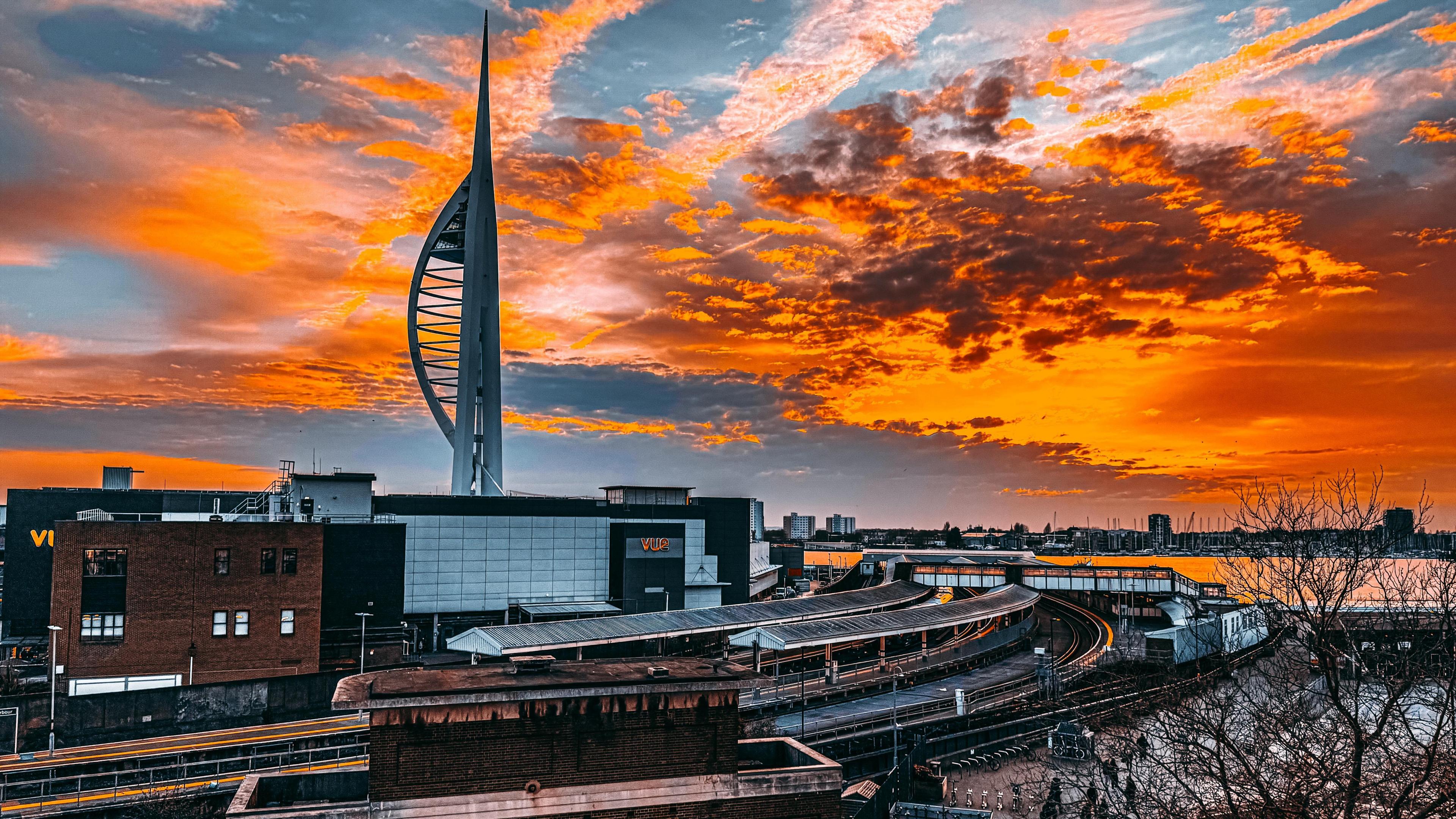 The sky glows a dramatic bright orange colour over Portsmouth. There are dark clouds on the horizon. Below you can see the Spinnaker Tower and train station. You can see the glass building of a cinema and beyond the water of Portsmouth Harbour.