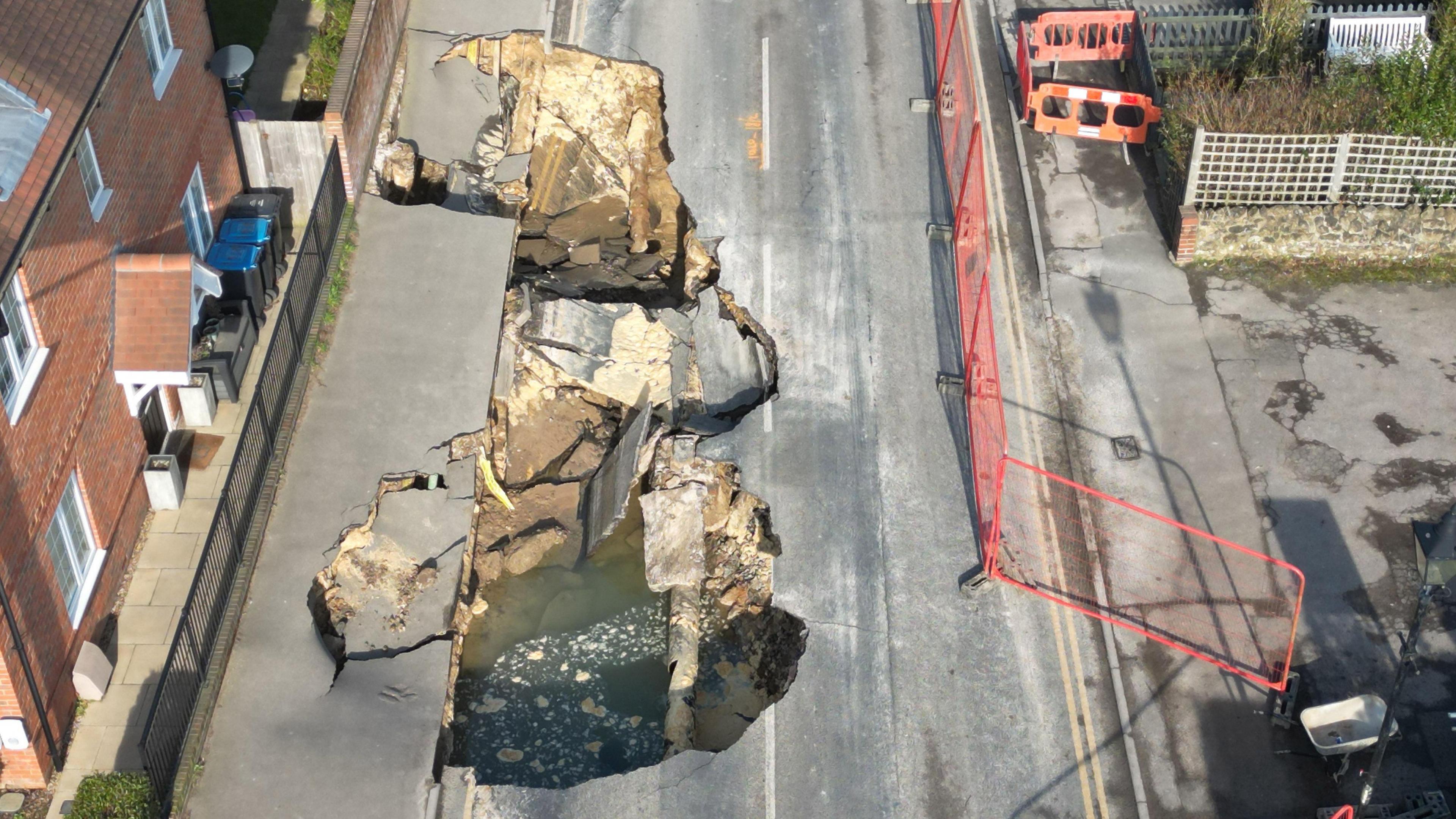 An aerial view of the sinkhole that has appeared in Godstone. One carriageway of the road has collapsed as well as two sections of pavement, with a damaged pipe and a pool of water visible in part of the sinkhole. A house is situated to the left, with the sinkhole now longer than its frontage.