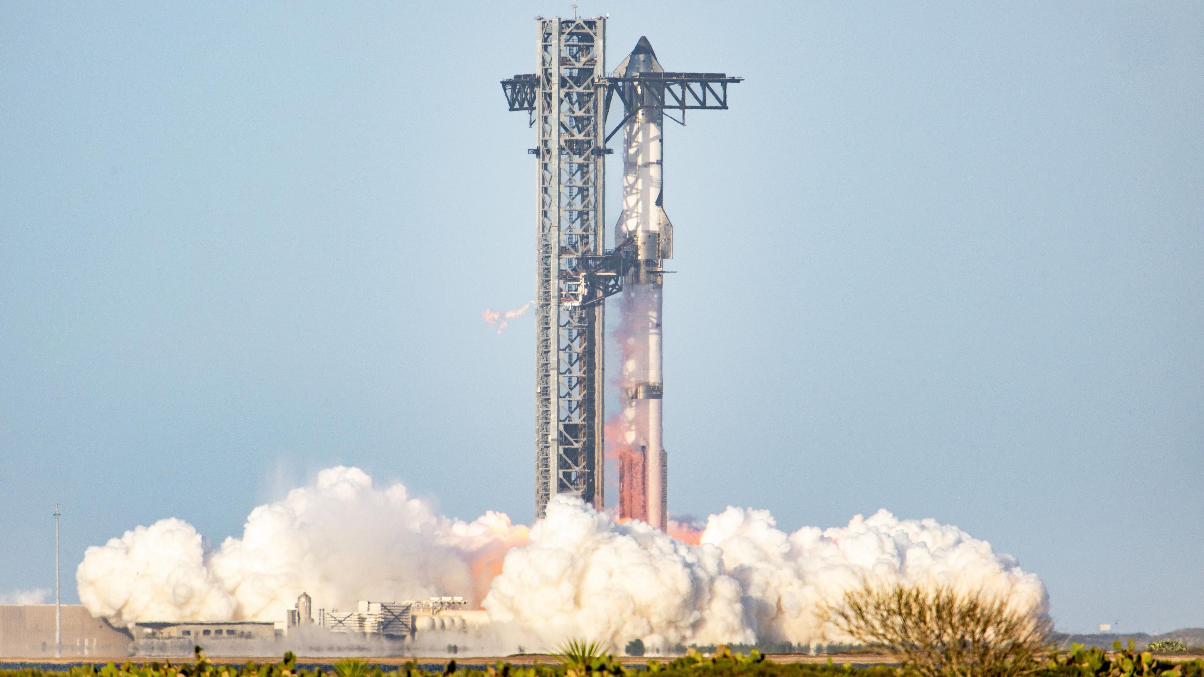 SpaceX Starship rocket takes off from a launch pad in Texas. There are blue skies and plumes of smoke can be seen rising from the ground. 