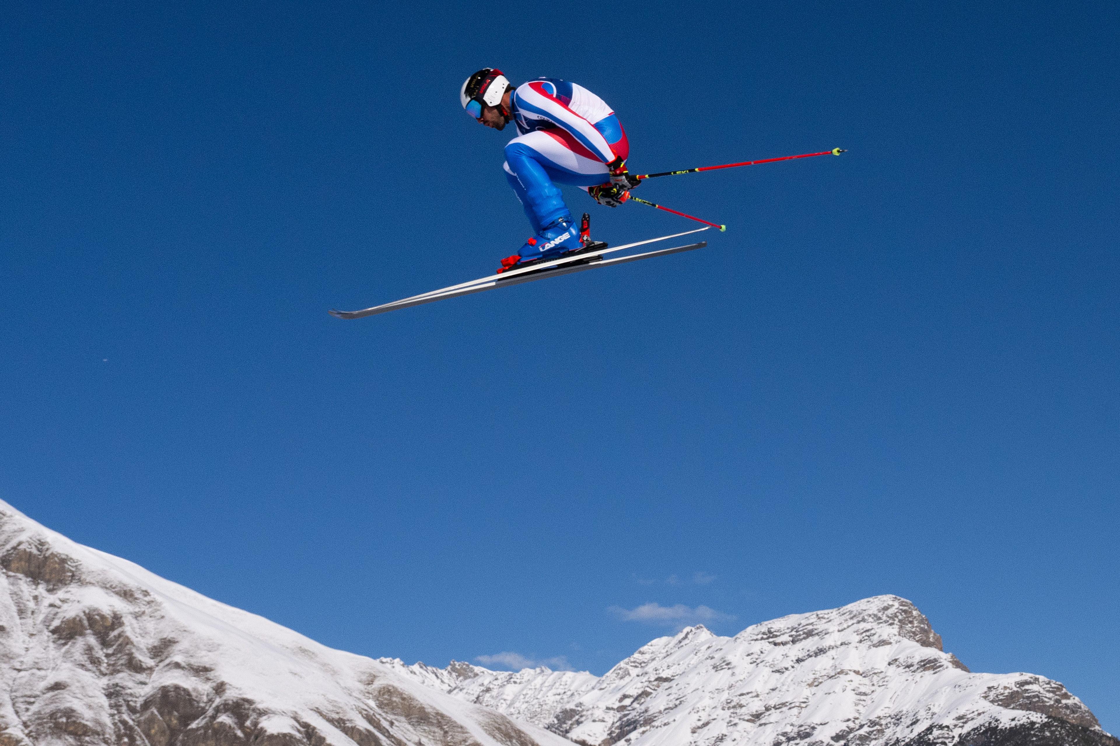 France's downhill skier Evan Klufts in a blue, white, and red racing suit soars high above a snowy mountain landscape, skis parallel and poles trailing behind, set against a clear blue sky.