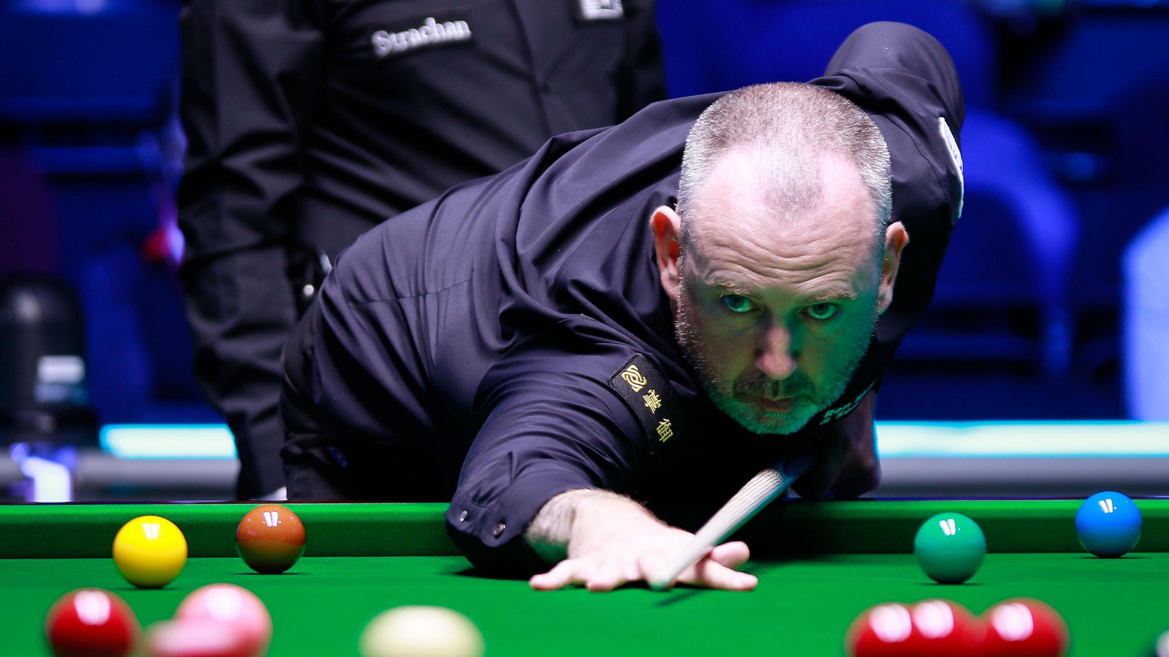 Mark Williams play a shot at a snooker table. He leans on the snooker table with one hand and holds the cue with another. He wears a black shirt and has short greying hair and stubble. 