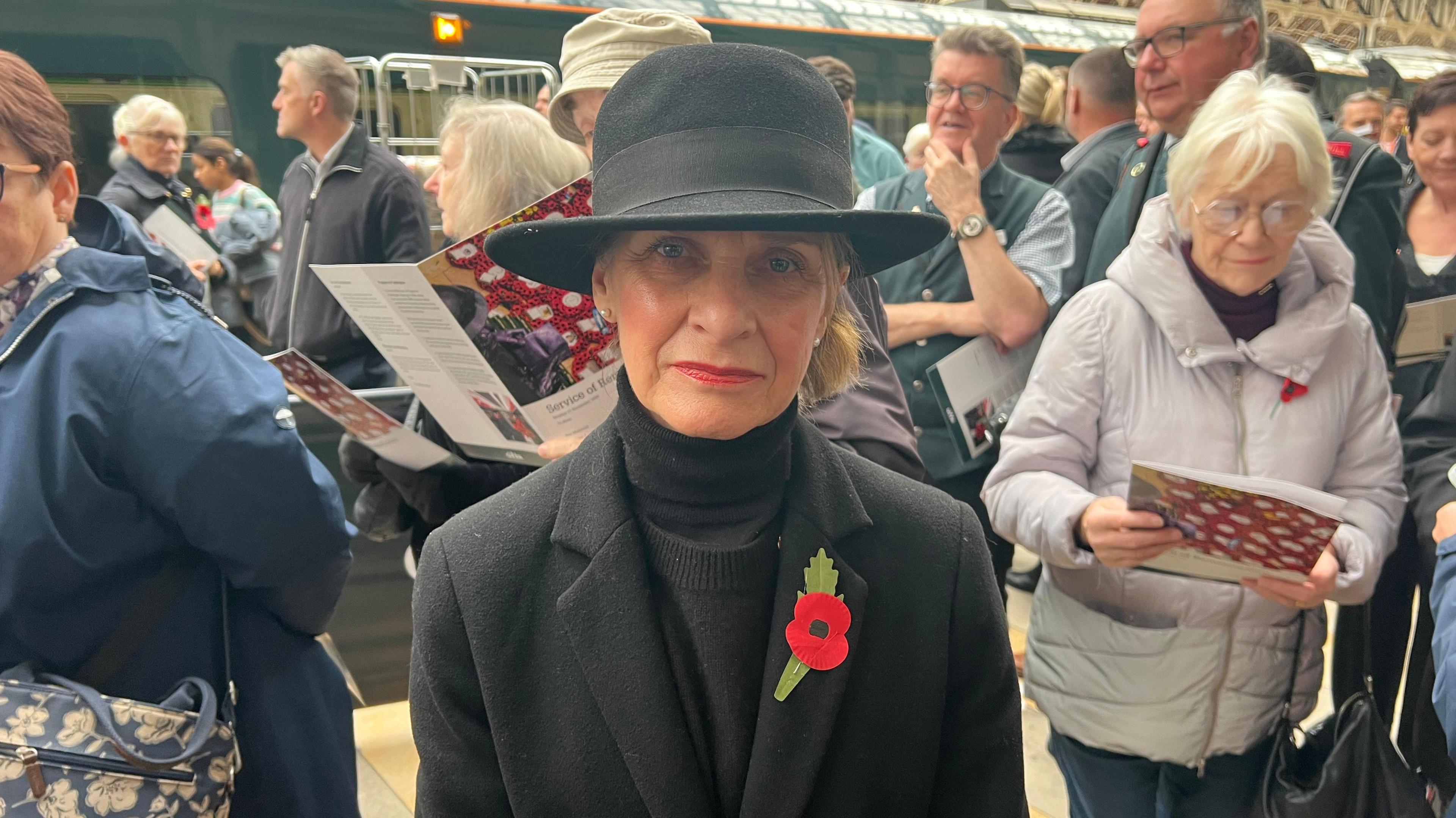 Wera Hobhouse, MP for Bath, dressed in a black hat, black turtleneck and a black blazer with a poppy on the lapel. She stands on a railway platform. There are lots of people in the background.