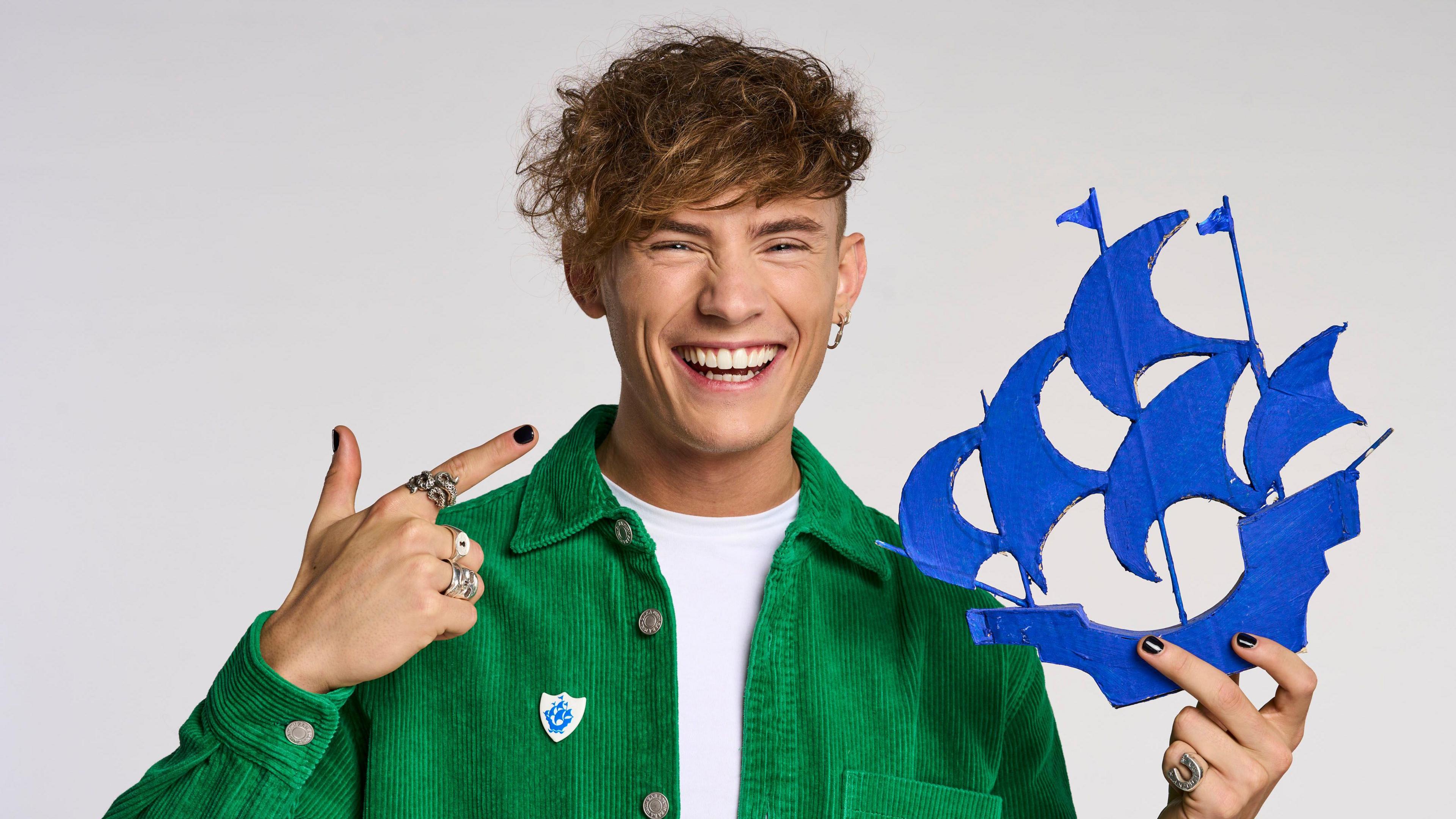 A man with dark curly hair is smiling and looking into the camera. He's dressed in a green shirt, white t-shirt and multiple rings. He's holding a cut out of the blue Blue Peter ship and point to it.