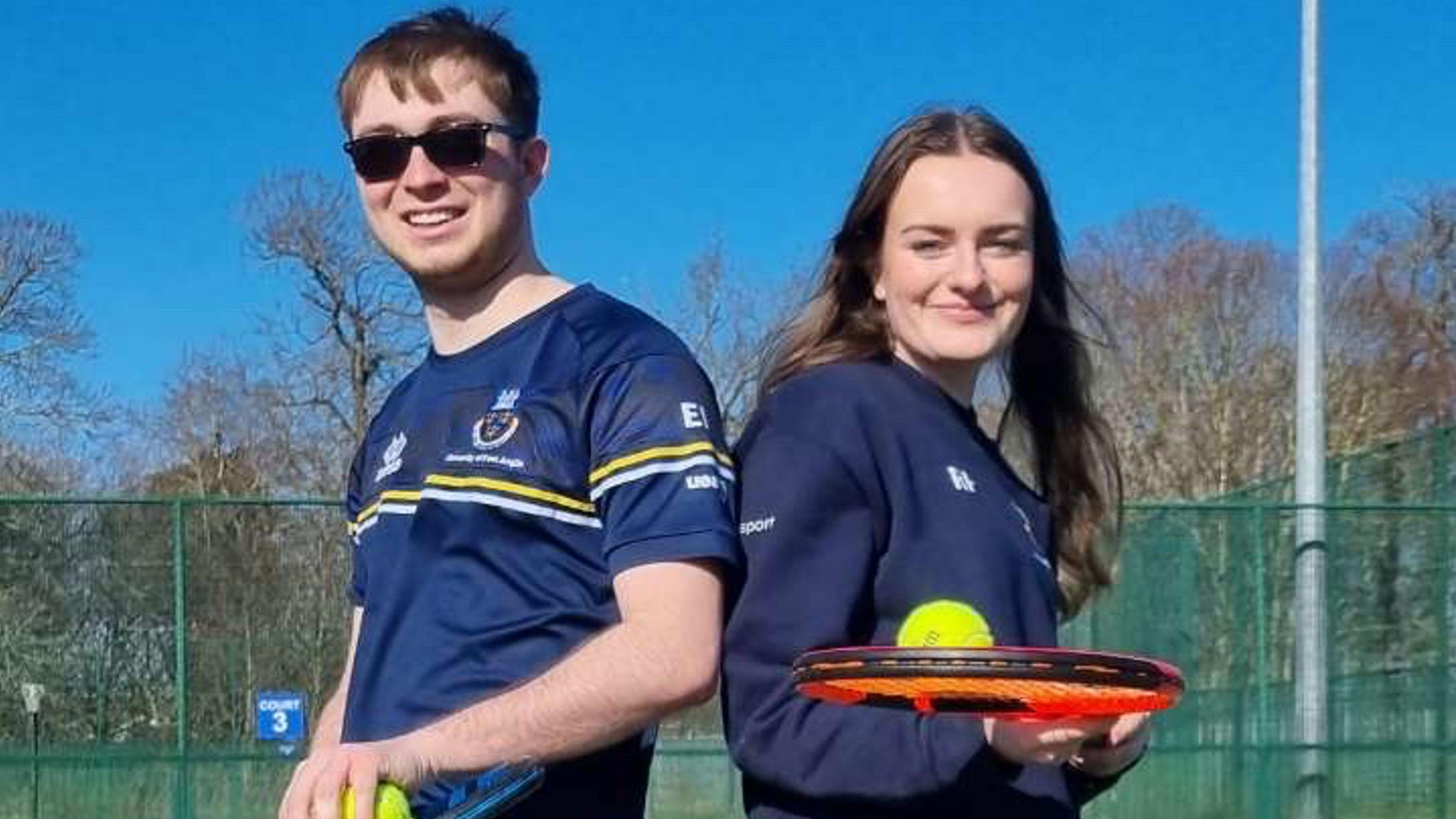 Ruby Pummell and Ed Ingles wearing blue tops and holding tennis rackets and balls. Ed has brown hair and is wearing sunglasses. Ruby has long brown hair. Both are smiling