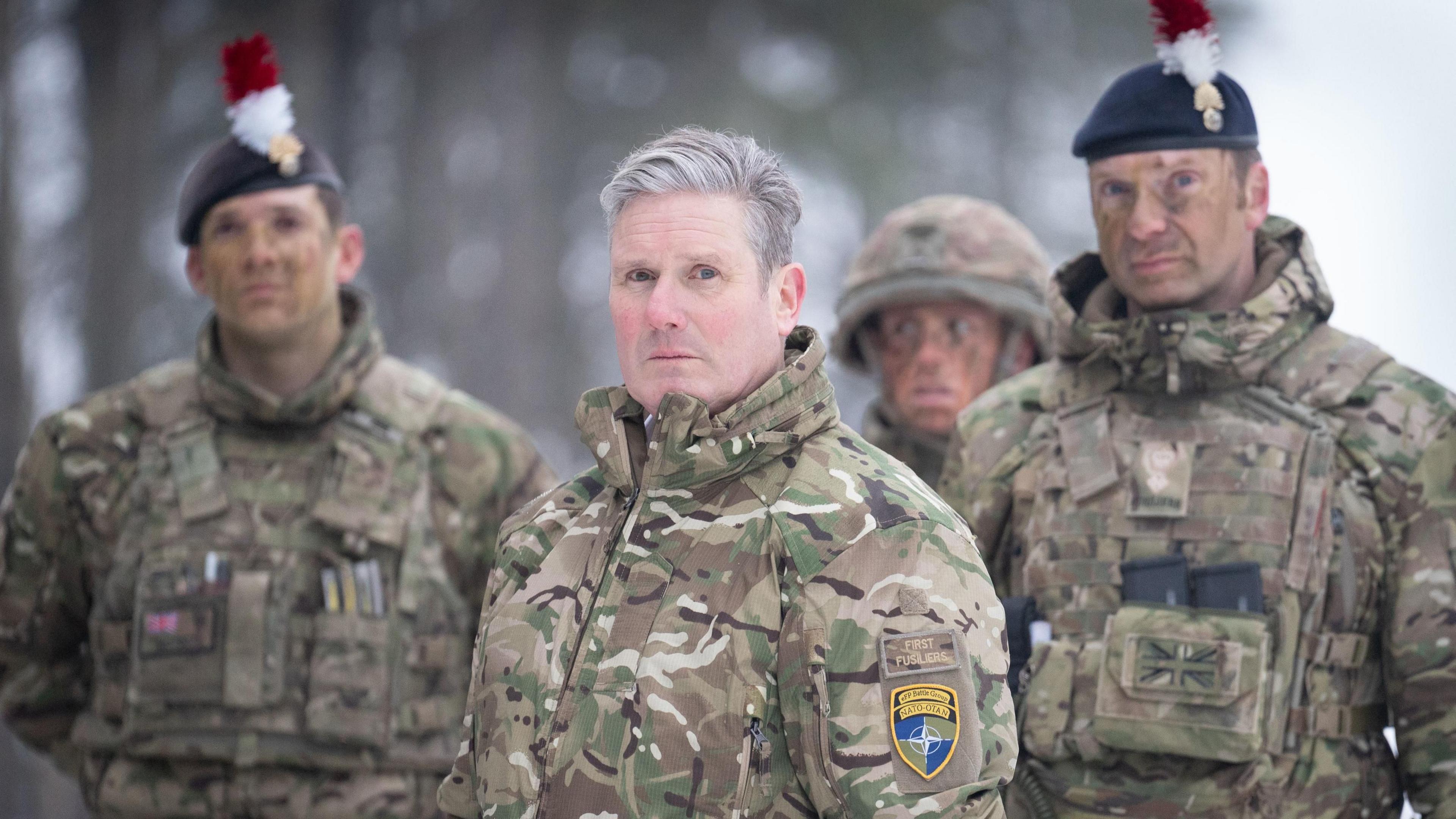 Sir Keir Starmer looks at the camera, as do two soldiers standing behind him, during his visit to Estonia. They are all wearing camouflaged jackets.