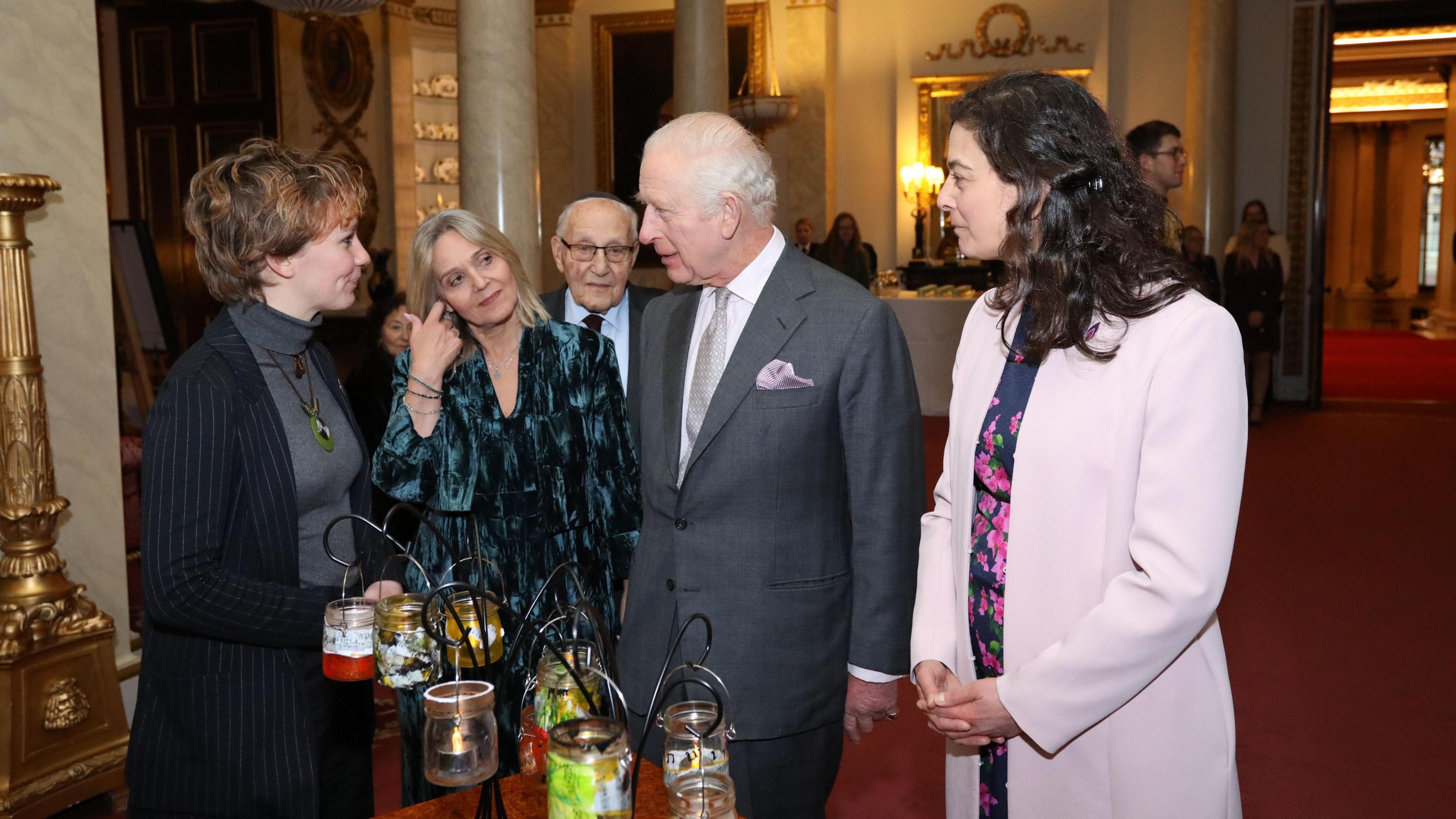 A woman with short brown hair and wearing a pinstripe blazer speaking to King Charles, who is wearing a grey suit. In front of them is a selection of glass candle jars suspended from black metal arms.