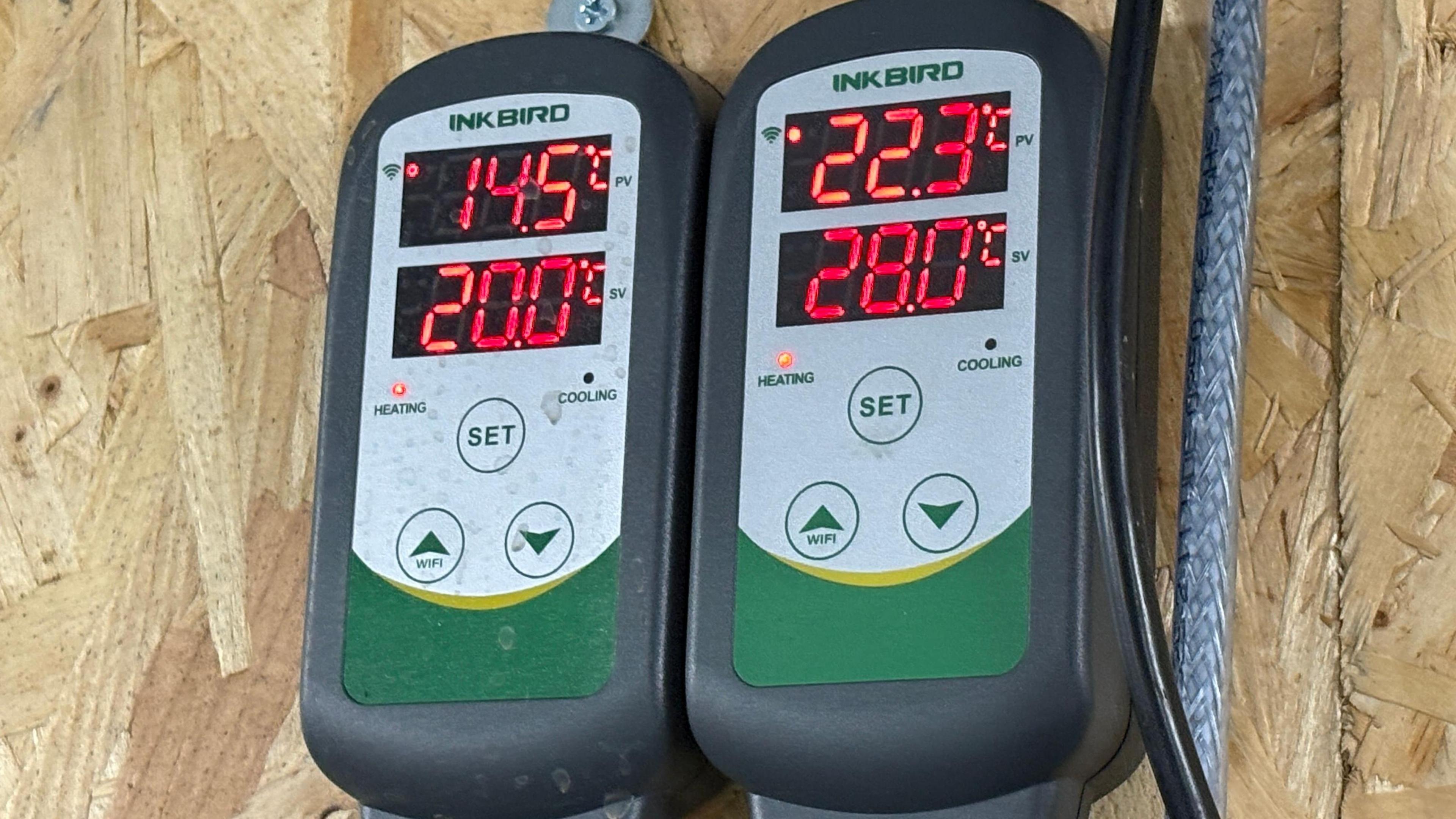 Two digital temperature gauges, mounted side by side on a piece of rough-looking wooden board. The gauge on the left shows two readings "14.5C" on the top and "20.0C" on the bottom. The gauge on the right reads "22.3C" on the top and "28.0C" on the bottom. The gauges bear the name "Inkbird" and give their readings on digital, red coloured numerical displays. Two wires or cables can be seen next to the gauge on the right.