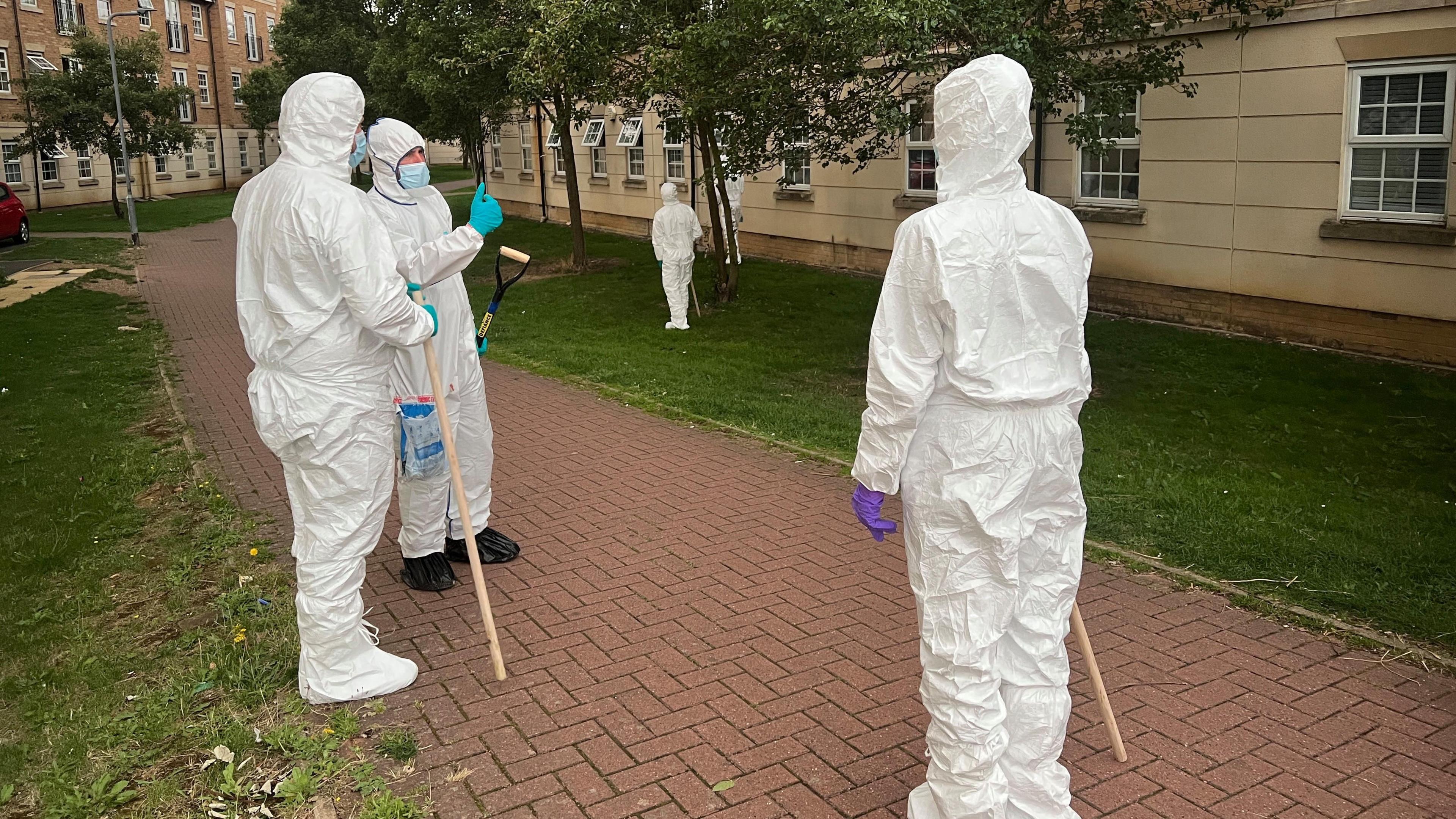 Three police officers wearing full-body white protective suits stand on a brick pathway bordered by grass. They are near blocks of flats with rows of windows. There are trees beside the path, which runs between the buildings. Two of the officers have poles they have been using to search the area. The third officer has a spade.