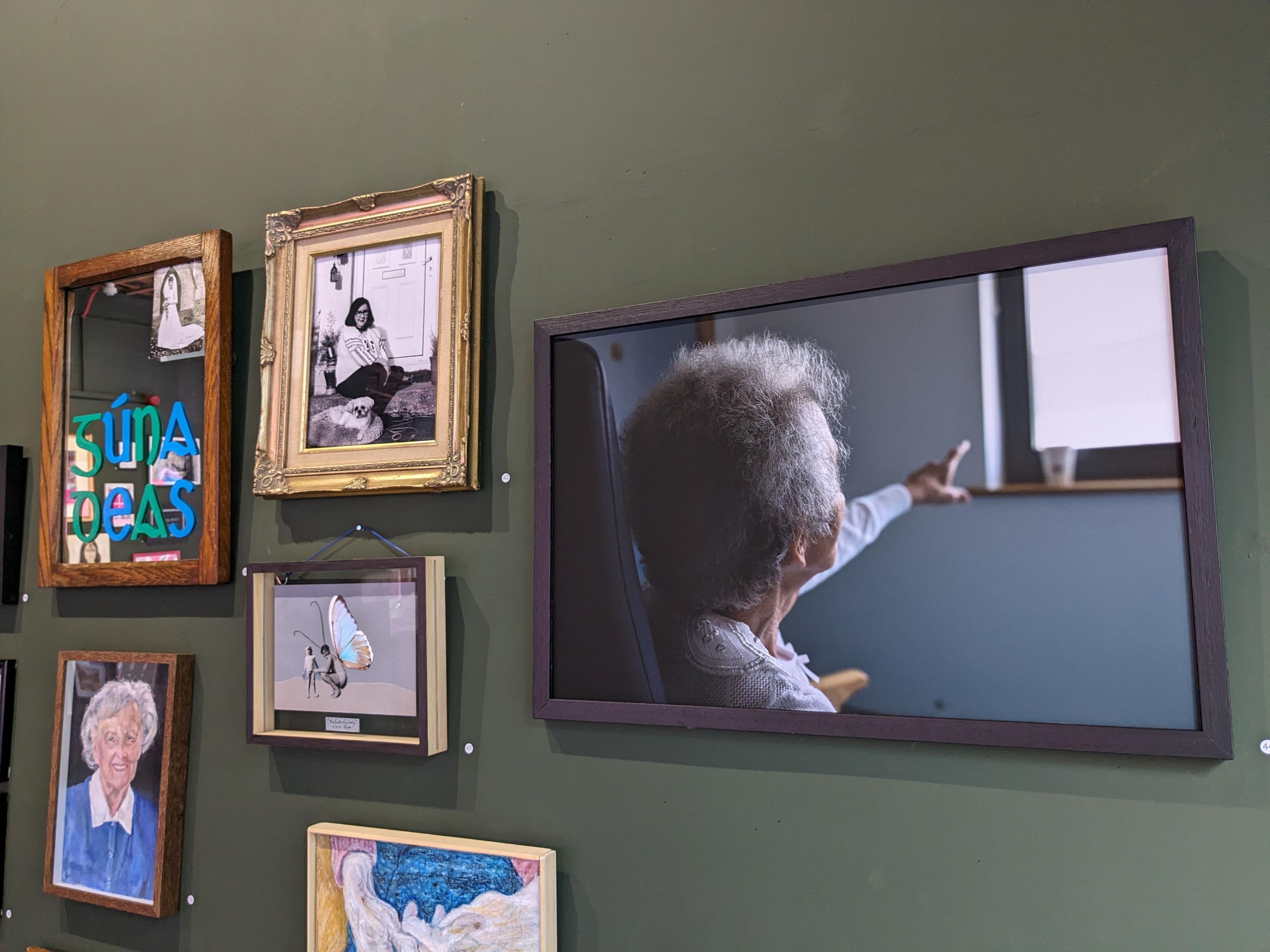 photograph of art on a wall; the most prominent piece depicts an older woman pointing out a window