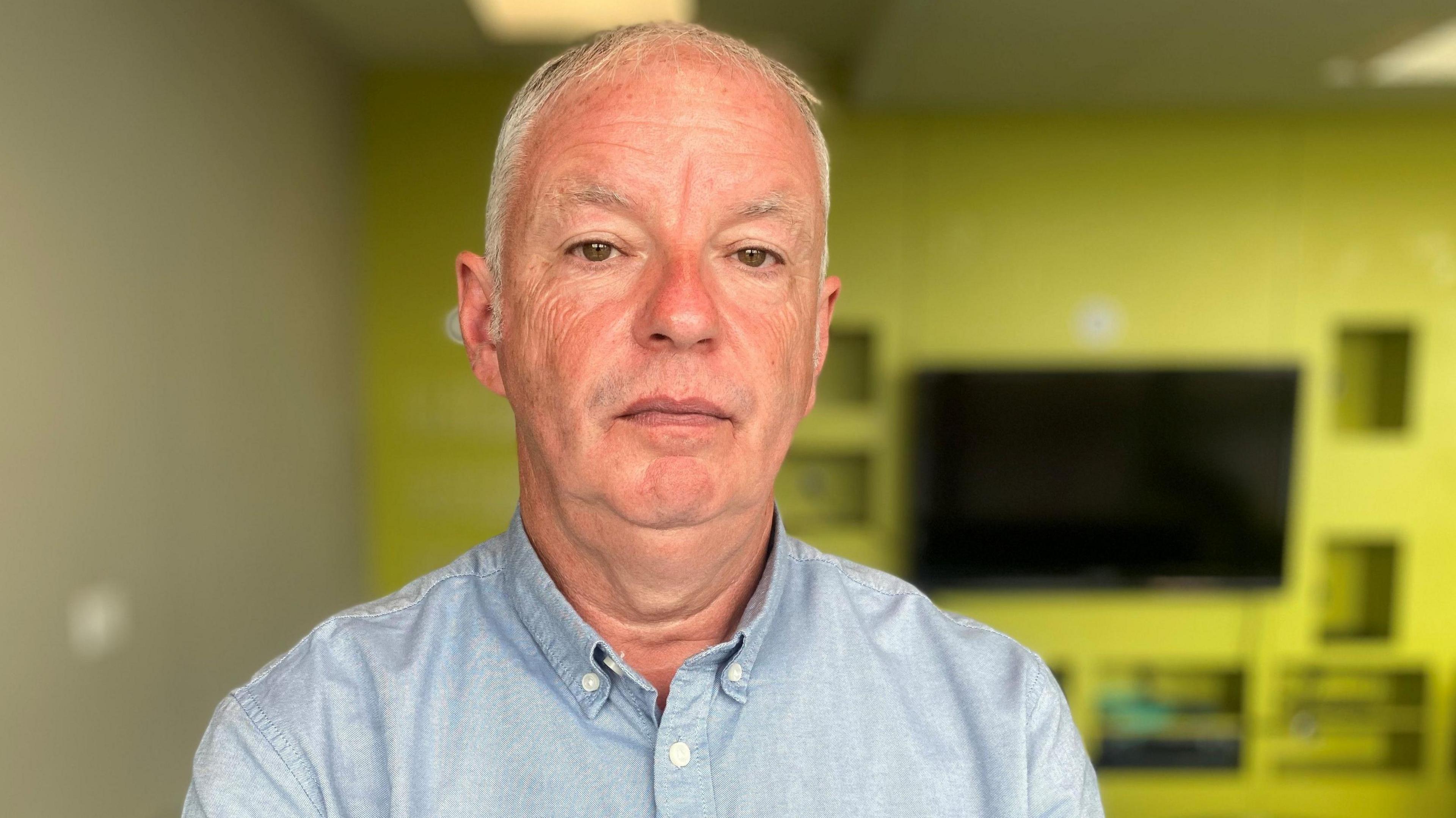 Gordon MacKenzie looking directly at the camera. He is wearing a blue shirt and has short grey hair. There is a yellow wall in the background with a wall-mounted TV and some cut-out sections for storage.