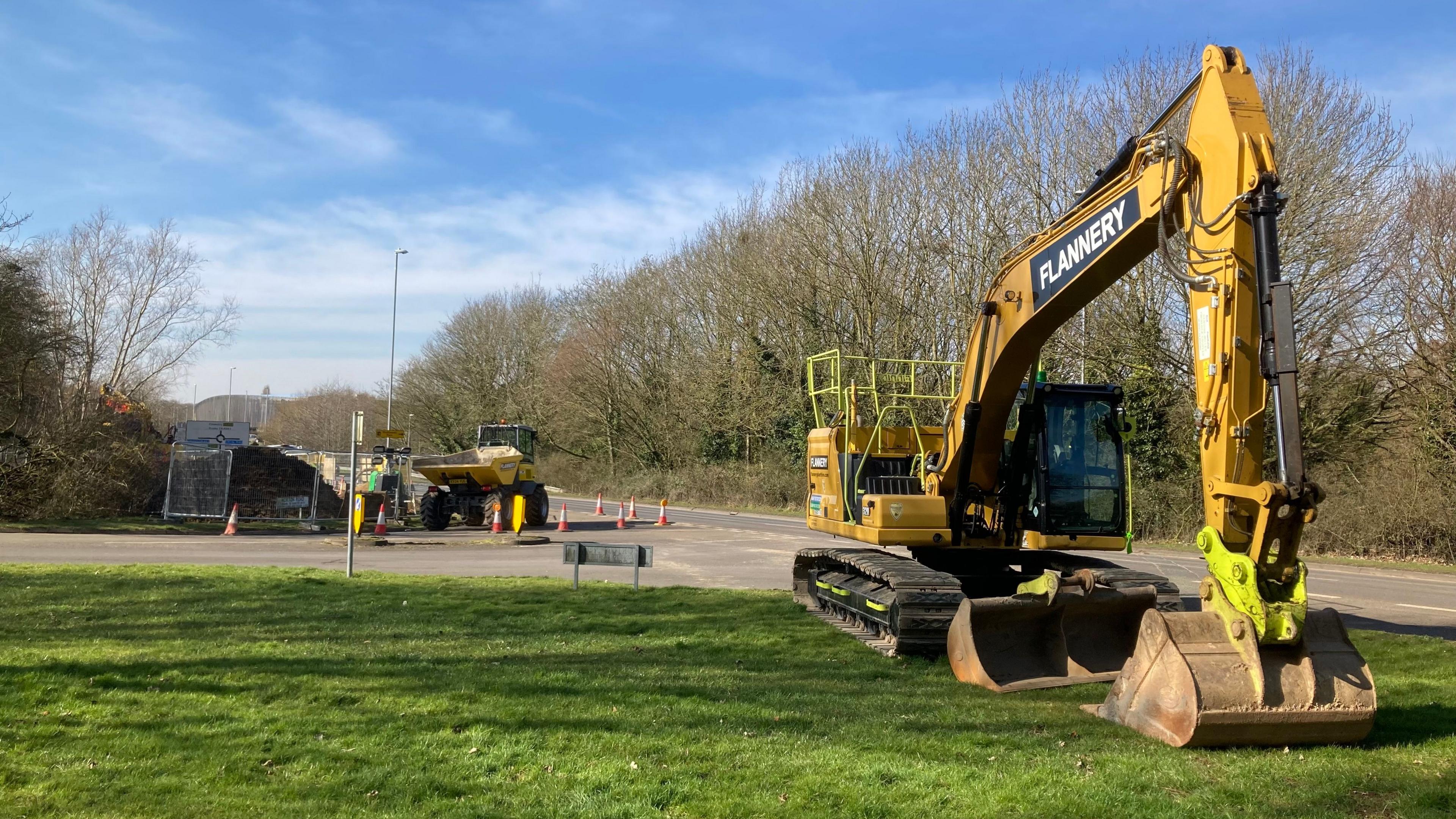 Yellow digger on grass close to the camera. Other plant visible in the background on the other side of a road junction. Three workers are just visible in the distance.