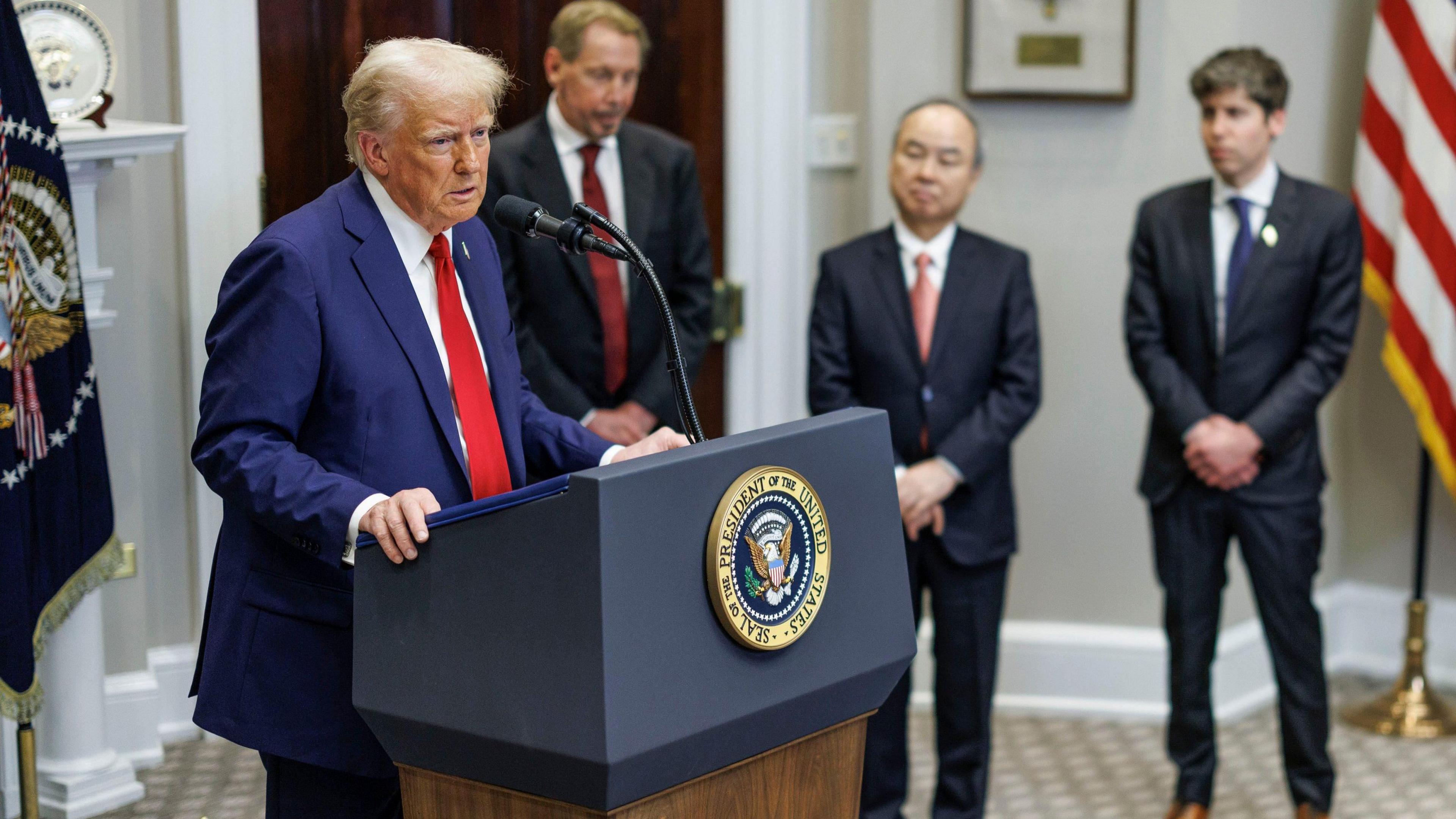 Donald Trump stands at a lectern in the White House, flanked by Larry Ellison, Masayoshi Son and Sam Altman.