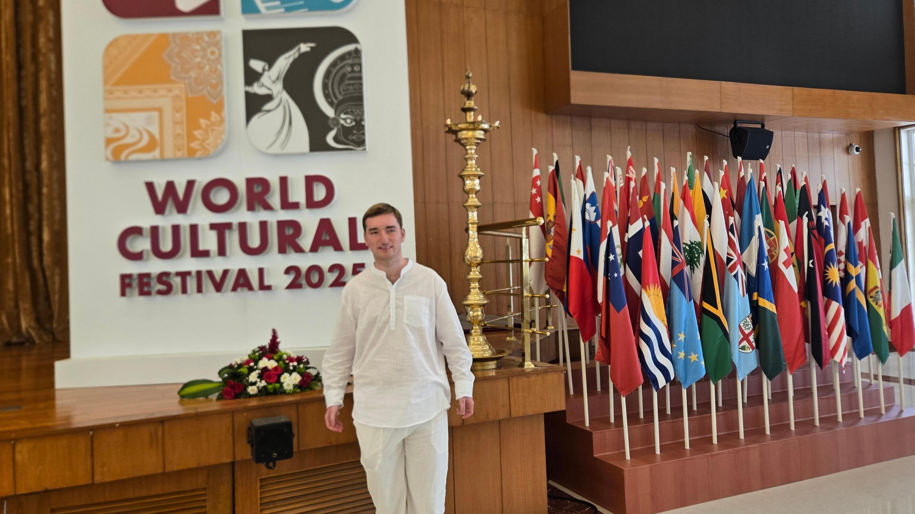 A man in white linen attire is smiling towards the camera and standing in front of a stage. Behind him is a board which reads World Cultural Festival 2025 and to his left there are a number of flags.