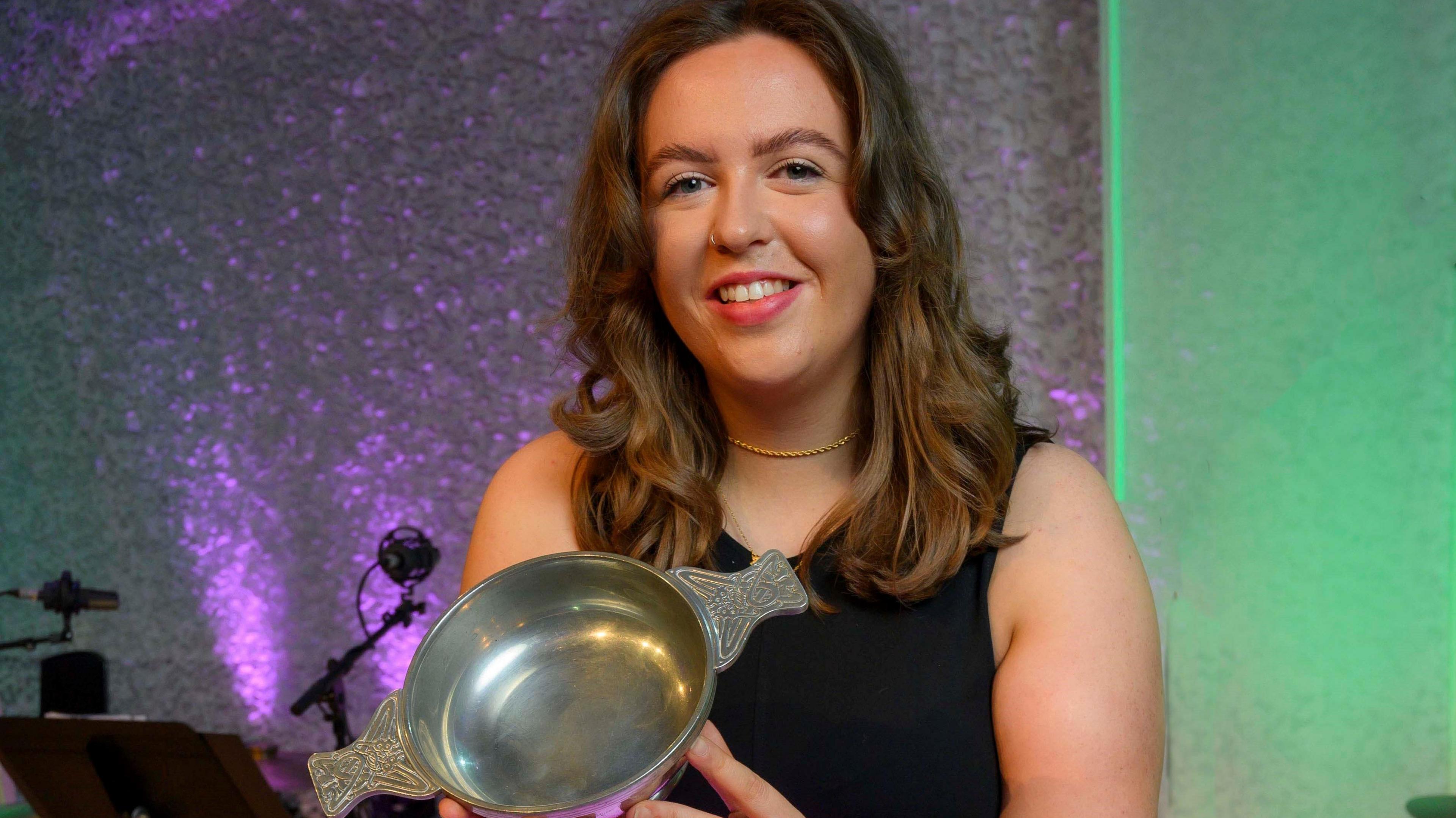 Ellie Beaton holds the hold the BBC Radio Scotland Young Traditional Musician quaich after winning the title for 2025. She has long waxy hear and is wearing a black sleeveless dress and a gold necklace. 