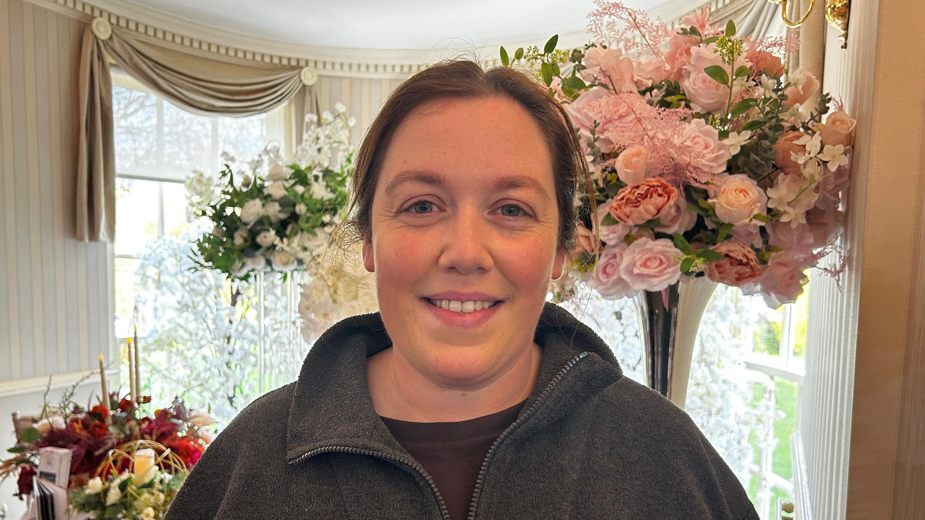 Mae Nia John yn edrych yn syth ymlaen ac yn gwenu. Mae ei gwallt wedi clymu nol ac mae'n gwisgo siwmper lwyd. Y tu ol iddi mae ffenest a blodau a rhosynod pinc. Mae hi'n ymweld â ffair briodas. 