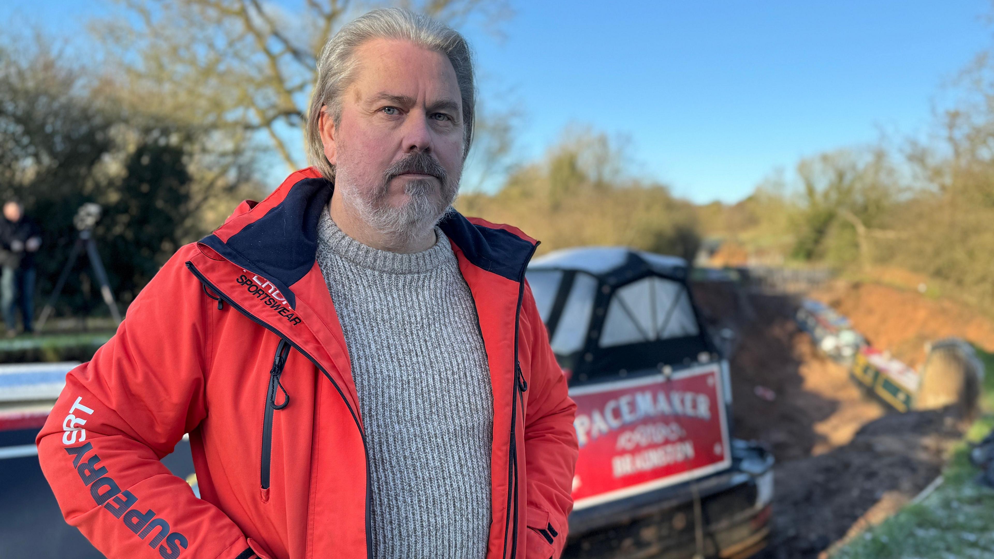A man with long swept back grey hair and grey facial hair, wearing a grey knit jumper and a red waterproof coat. He is standing in front of a dried out section of canal and there is a black and red canal boat behind him called the Pacemaker